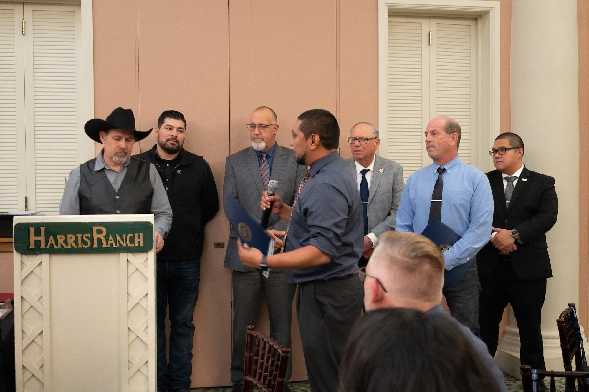 A group of men at an indoor event, one speaking at a podium labeled "Harris Ranch." Some hold certificates or wear formal attire.