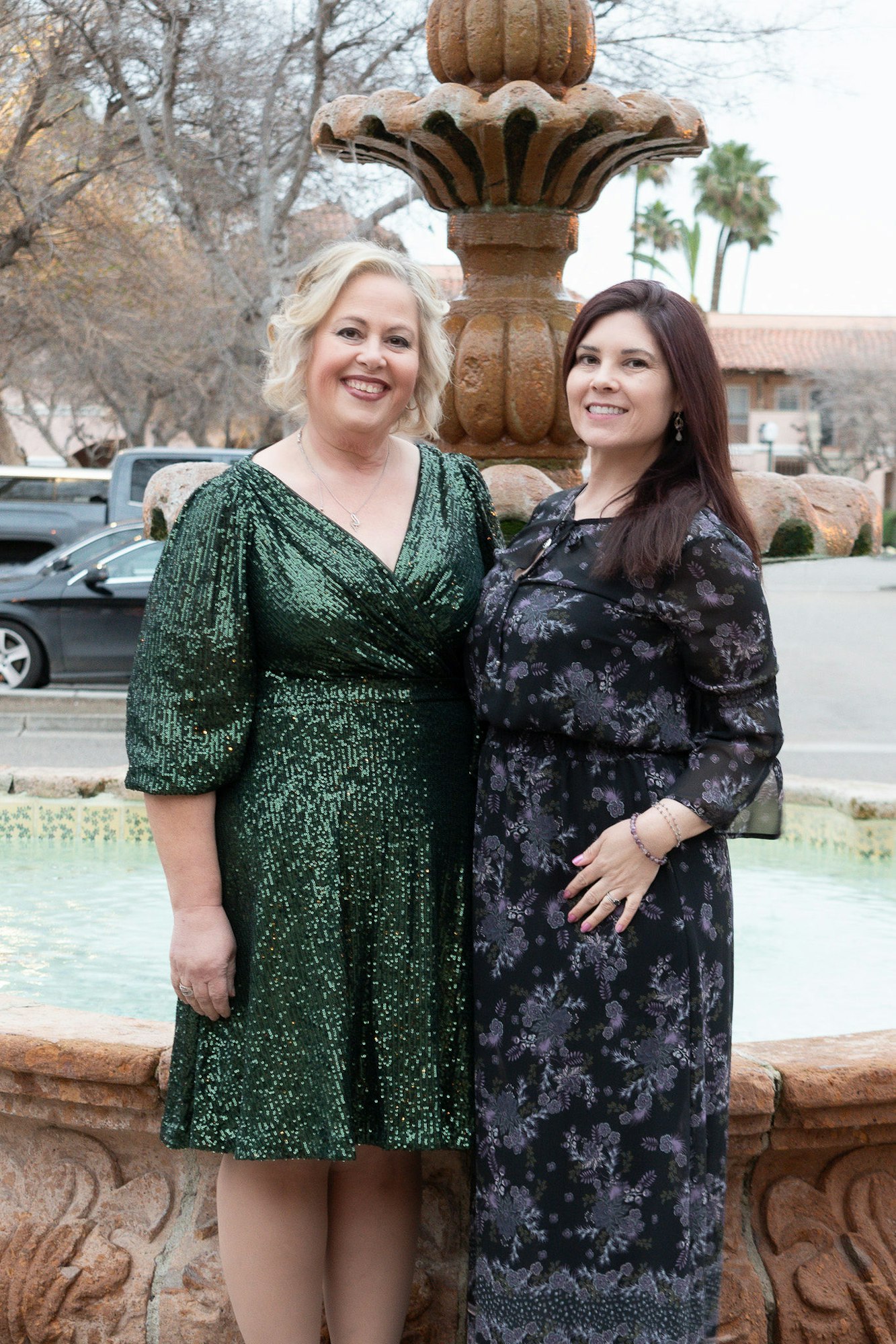 Two smiling women in dresses standing in front of a fountain with trees and a building in the background.
