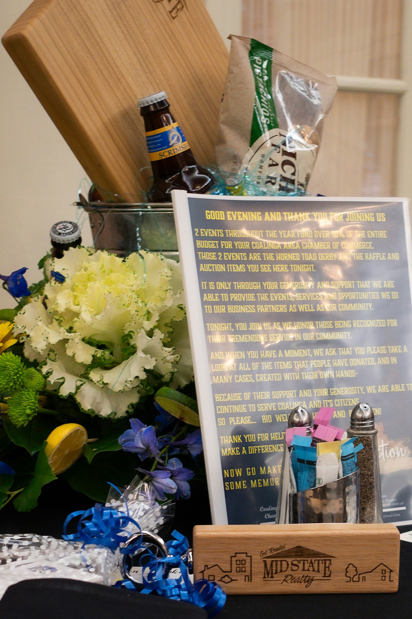 A gift basket with various items, a wooden board, bottle, snacks, and a sign about thanking and bidding, likely at a charity event.