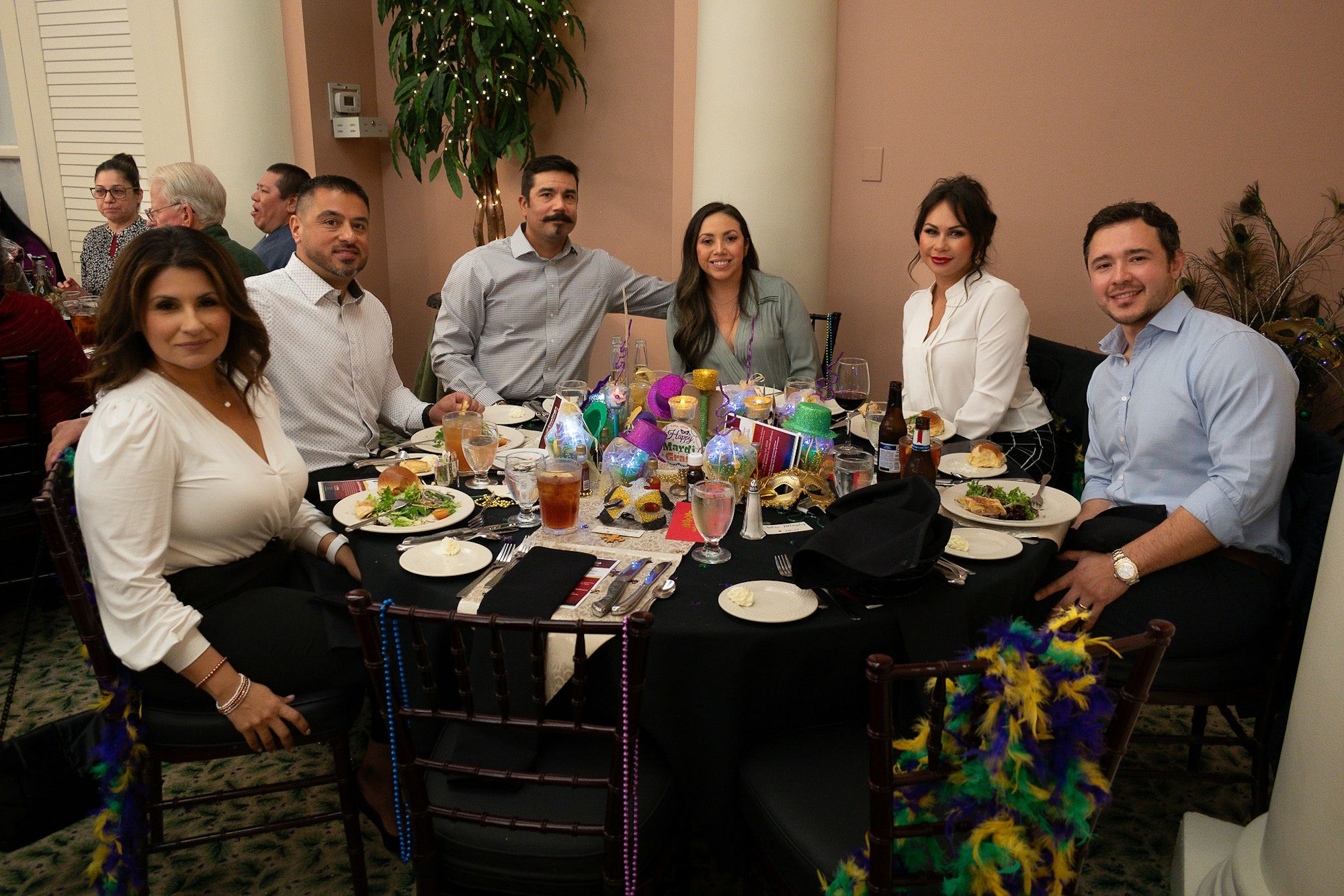 Group of people smiling at a festive table with masks and beads, suggesting a Mardi Gras themed event.