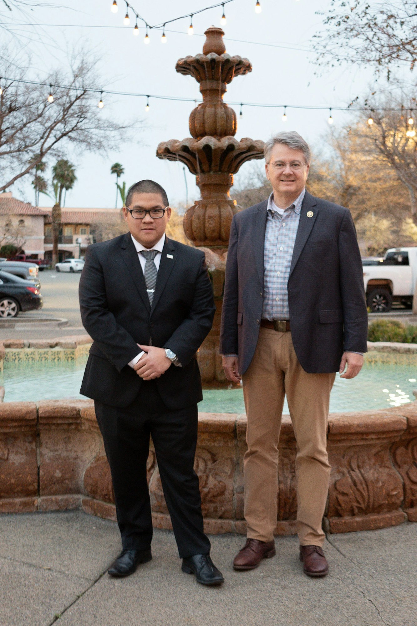 Two men in suits standing by a fountain with string lights overhead.