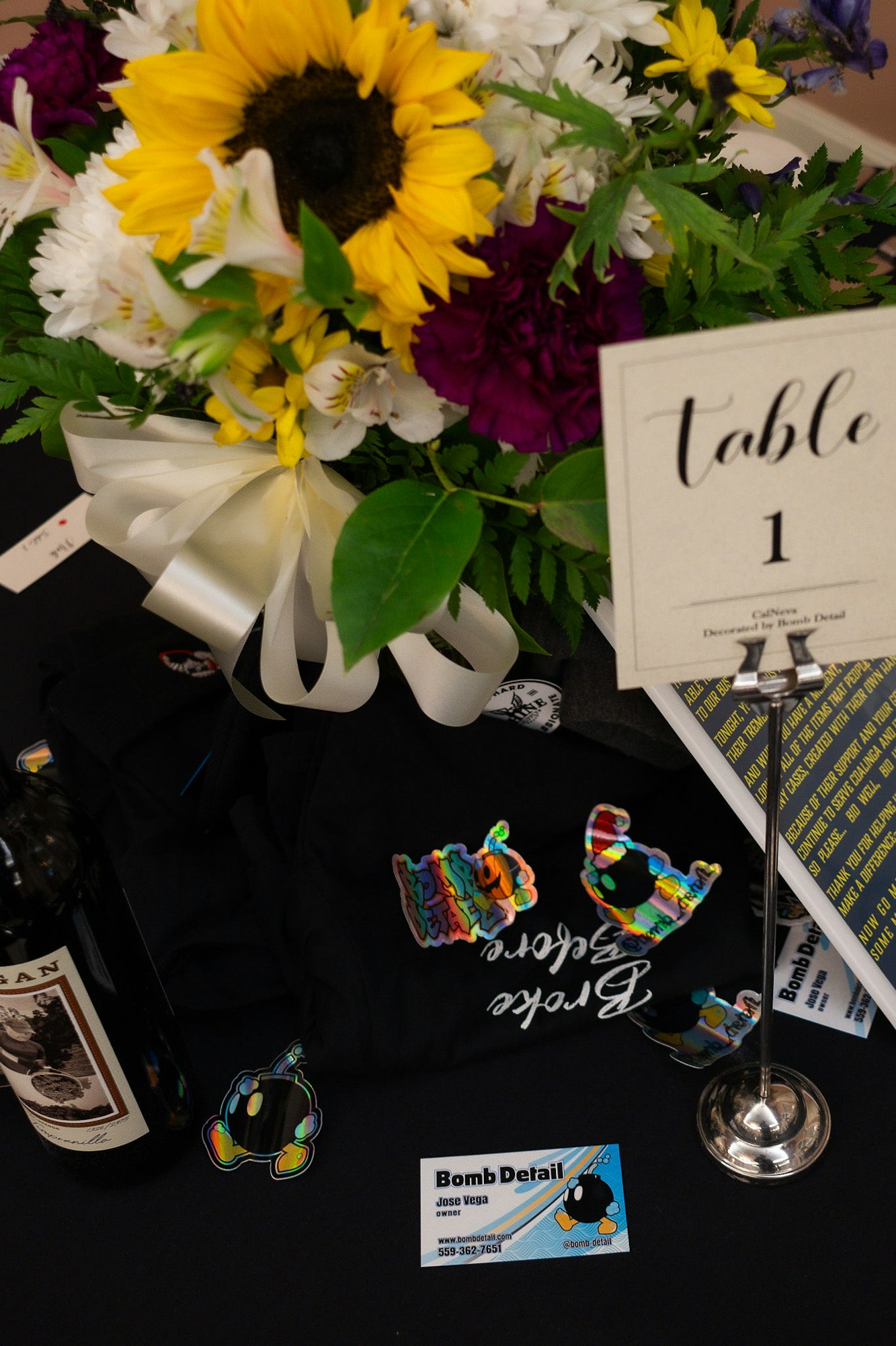 A table display with a bouquet, "Table 1" sign, a wine bottle, a T-shirt, and business cards.