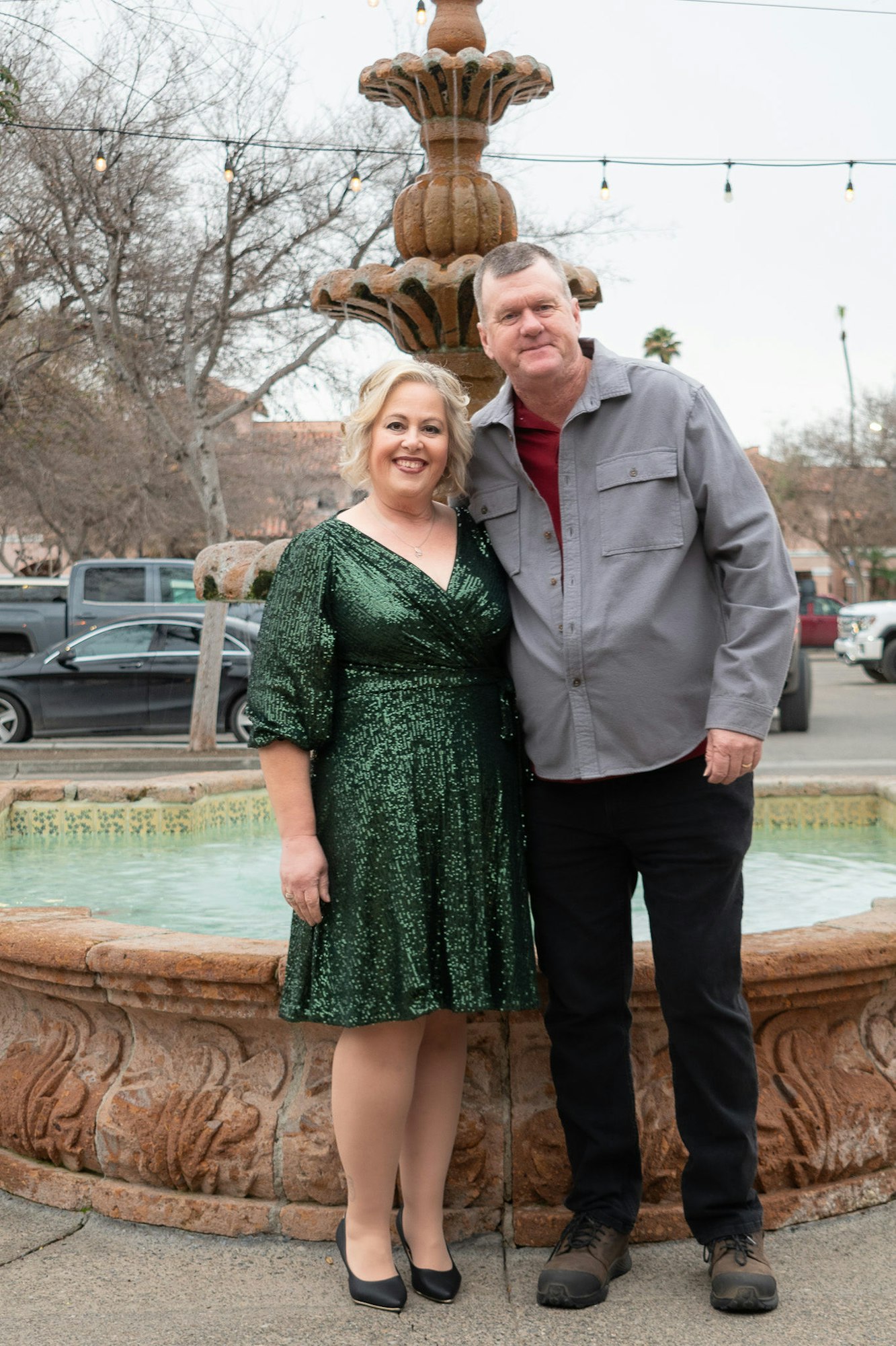 Two people posing in front of a fountain with hanging lights in the background.