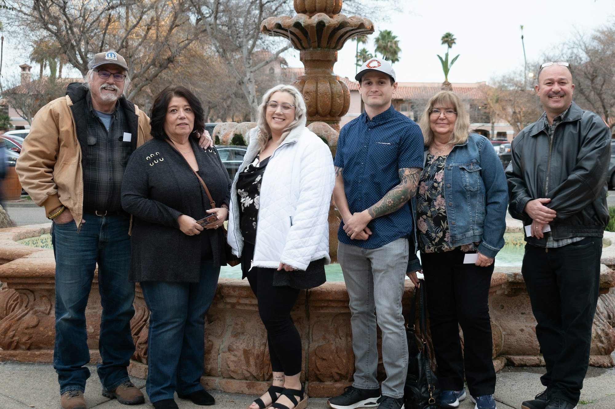 Group of six adults posing in front of a fountain, smiling, possibly a family gathering.