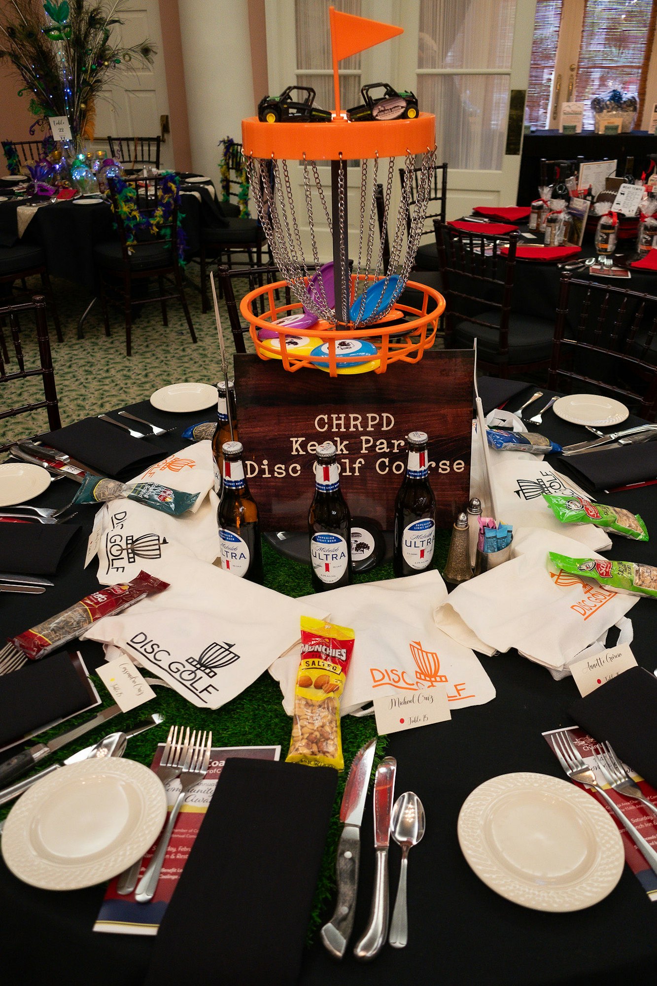 A disc golf basket centerpiece on a decorated table with snacks, beverages, and themed accessories for a disc golf event.