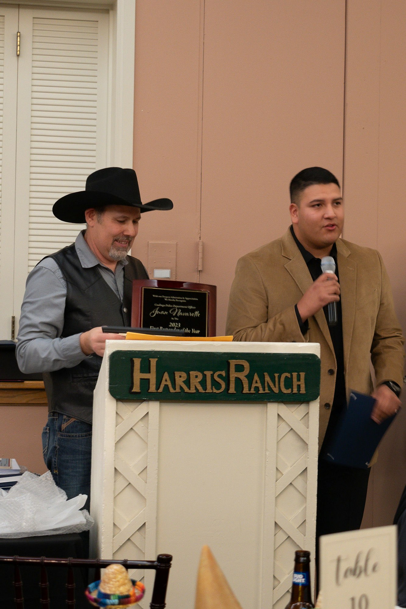 Two men at a podium with a "Harris Ranch" sign, one holding an award, the other speaking into a microphone.