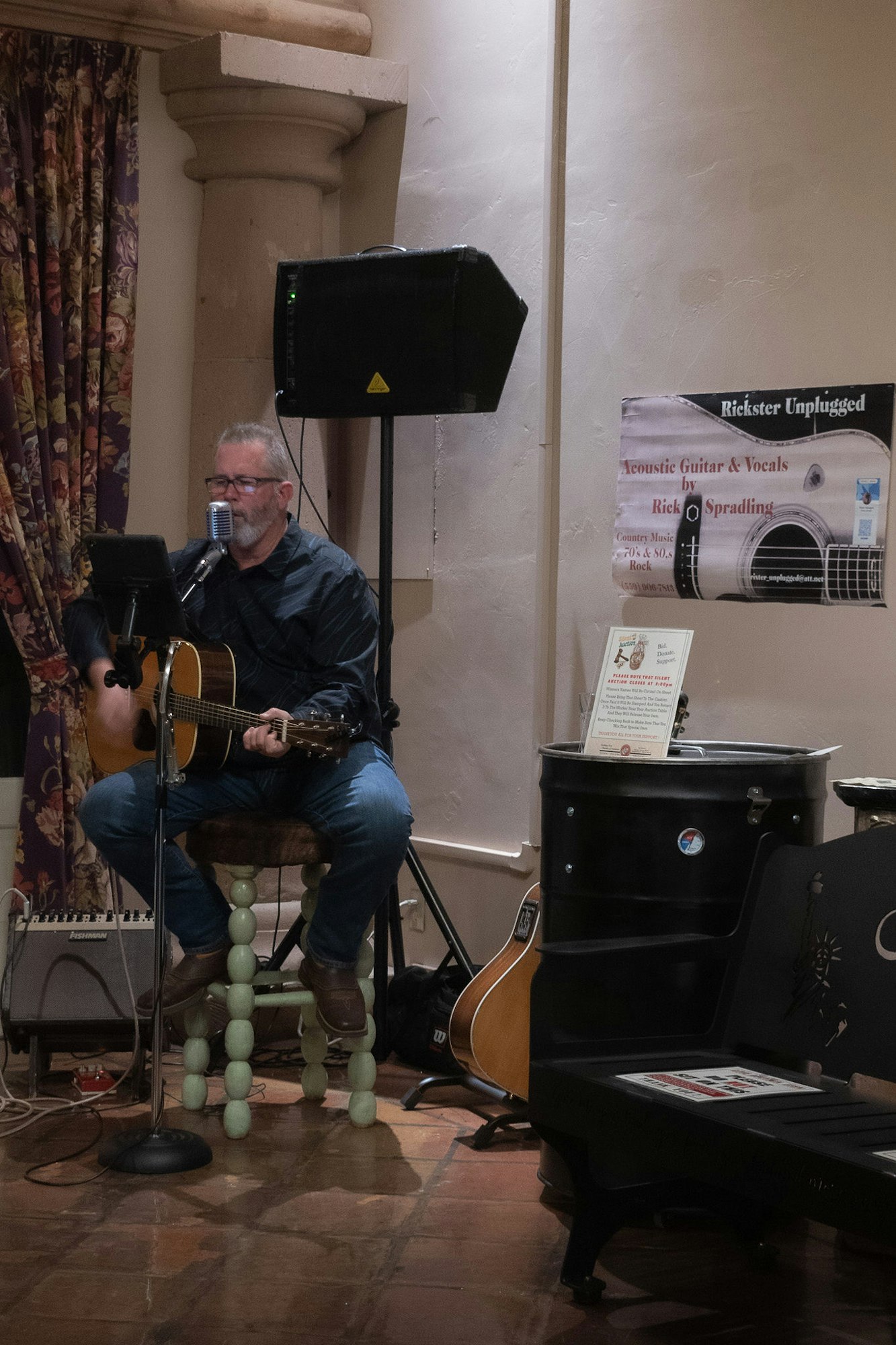 A musician playing guitar & singing into a microphone, with a speaker, promo banner, guitar cases, and an amp nearby.