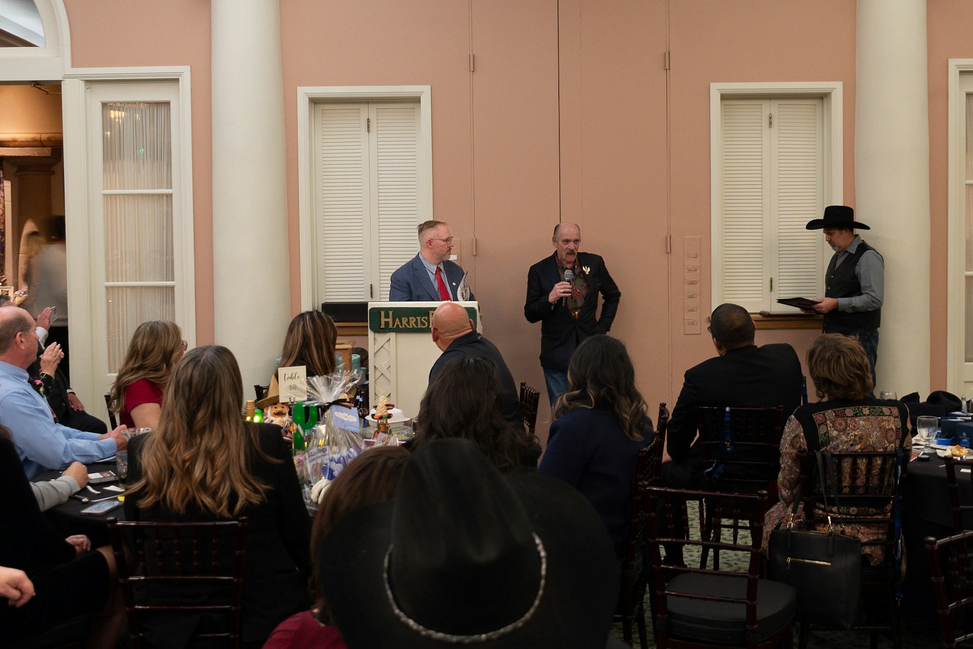 People at an indoor event with a speaker at a podium and another holding a microphone, audience listening, some wearing cowboy hats.