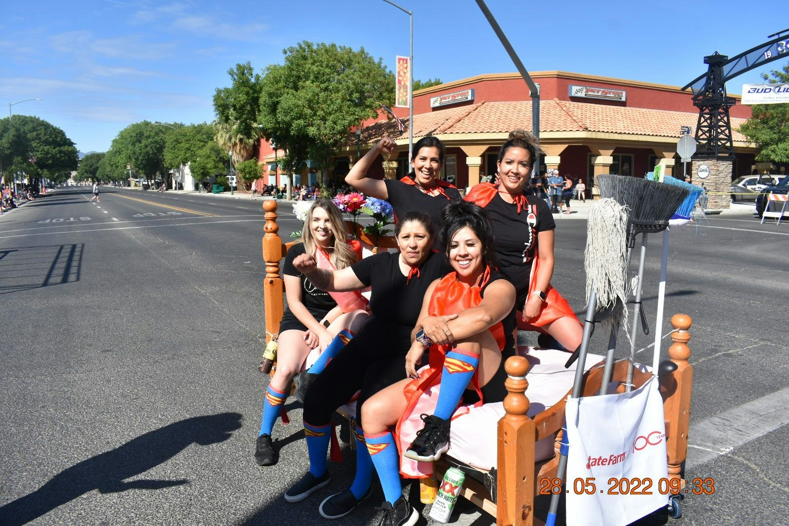 Group of five women in superhero attire sitting on a bed in the street, laughing and posing.