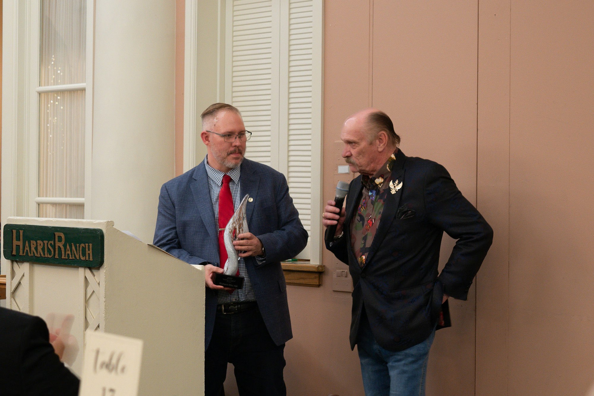 Two men at an event, one with a trophy and the other with a microphone, near a sign that reads "Harris Ranch."