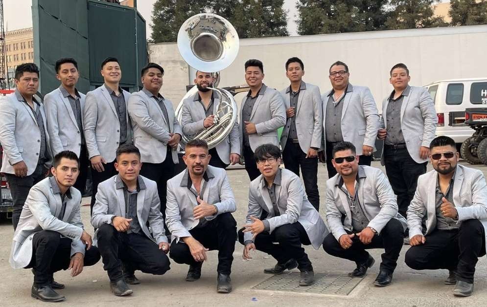 A group of men in matching silver jackets poses together, with one holding a tuba, likely a musical band or ensemble.