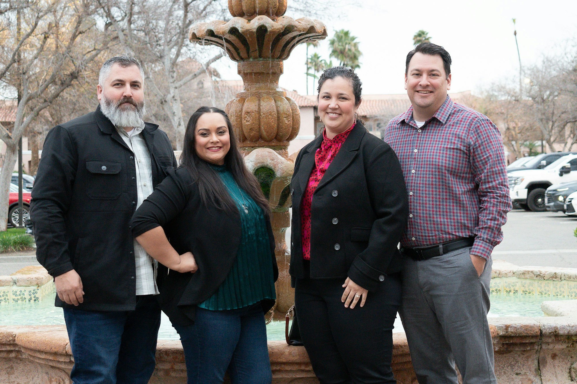 Four people posing in front of a water fountain, smiling, dressed in semi-formal attire.