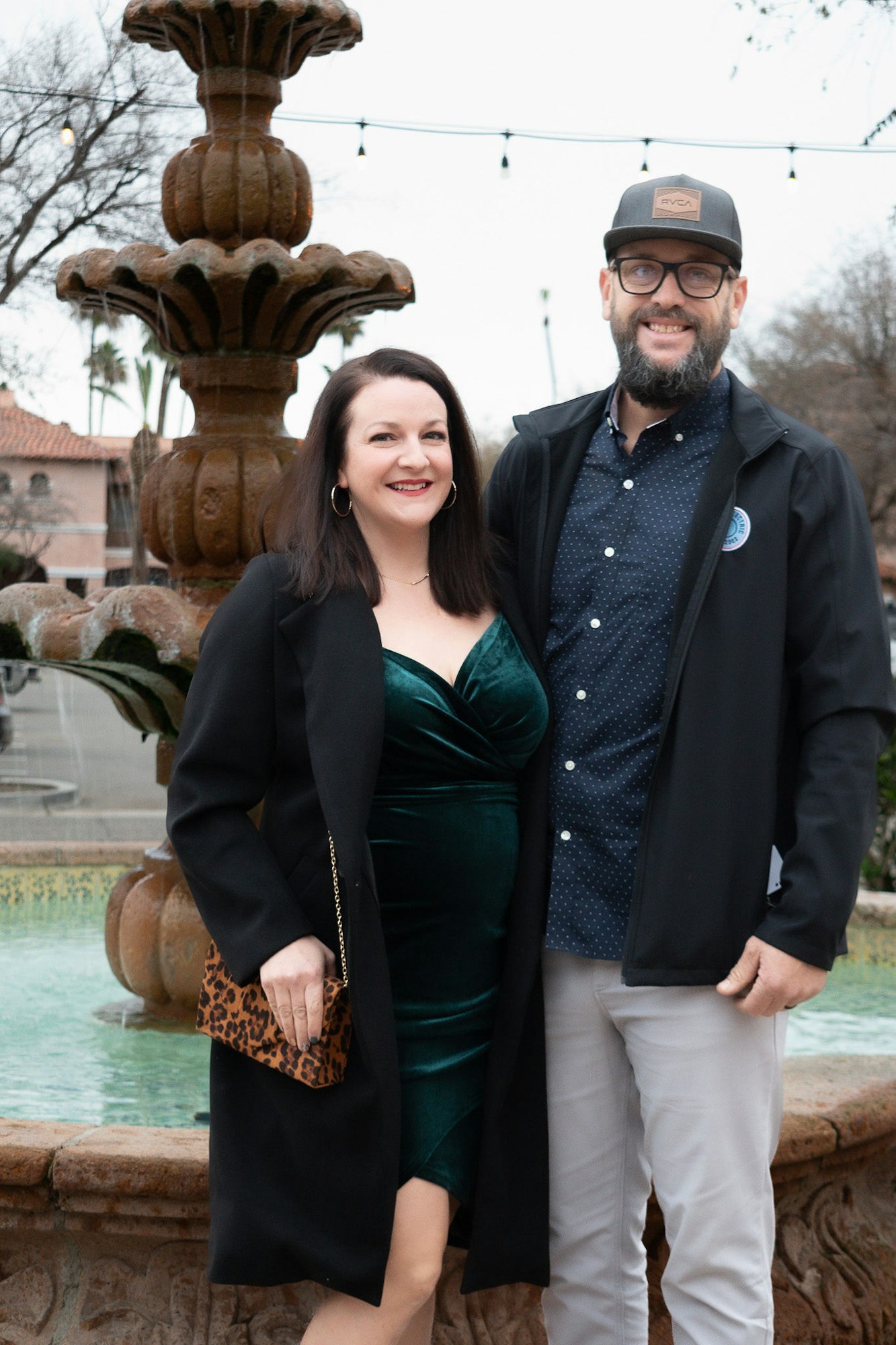 A smiling couple posing in front of a fountain, dressed elegantly for an event.