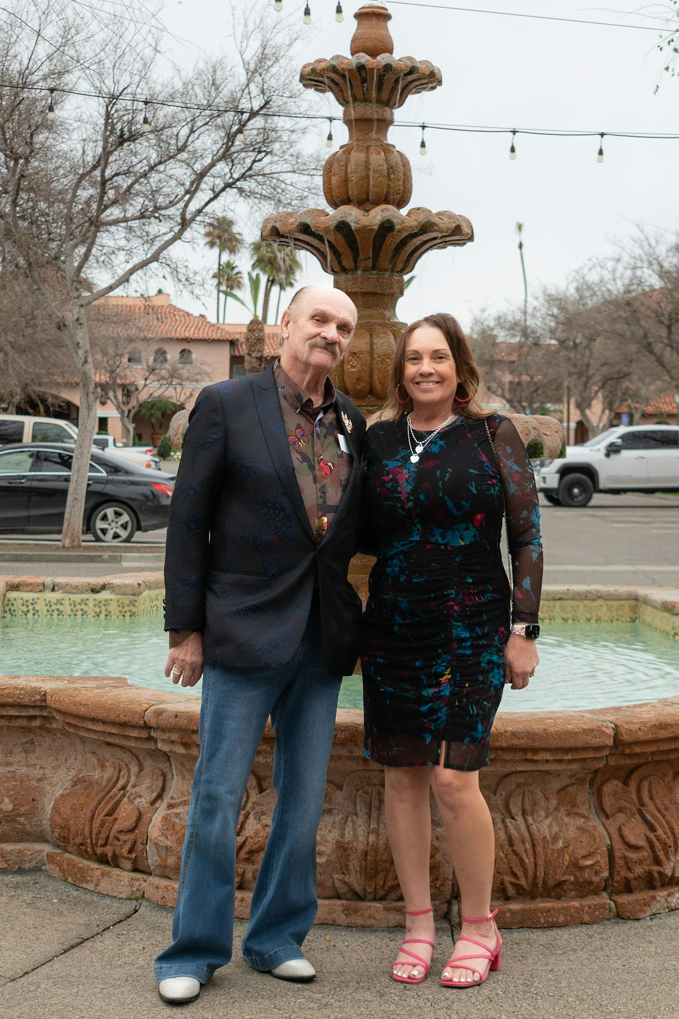 Two people smiling in front of a fountain, dressed in smart casual attire.