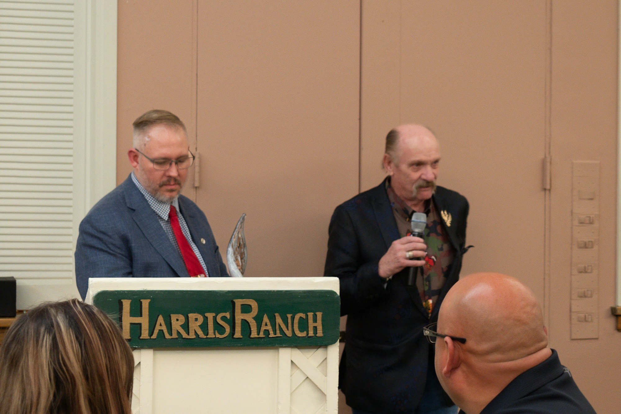 Two men at a podium with a "Harris Ranch" sign, one with a mic speaking, the other holding an award, in a room with listeners.