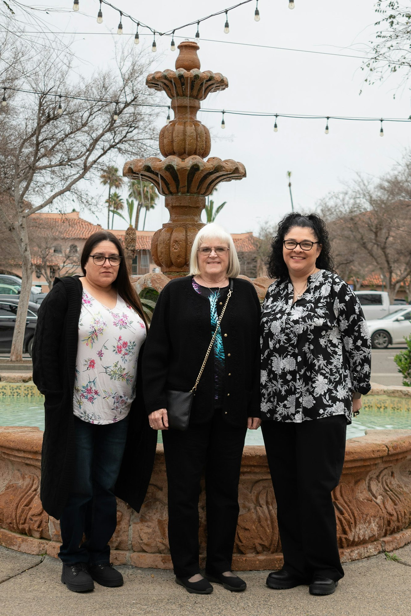 Three people posing in front of a fountain with hanging lights above.