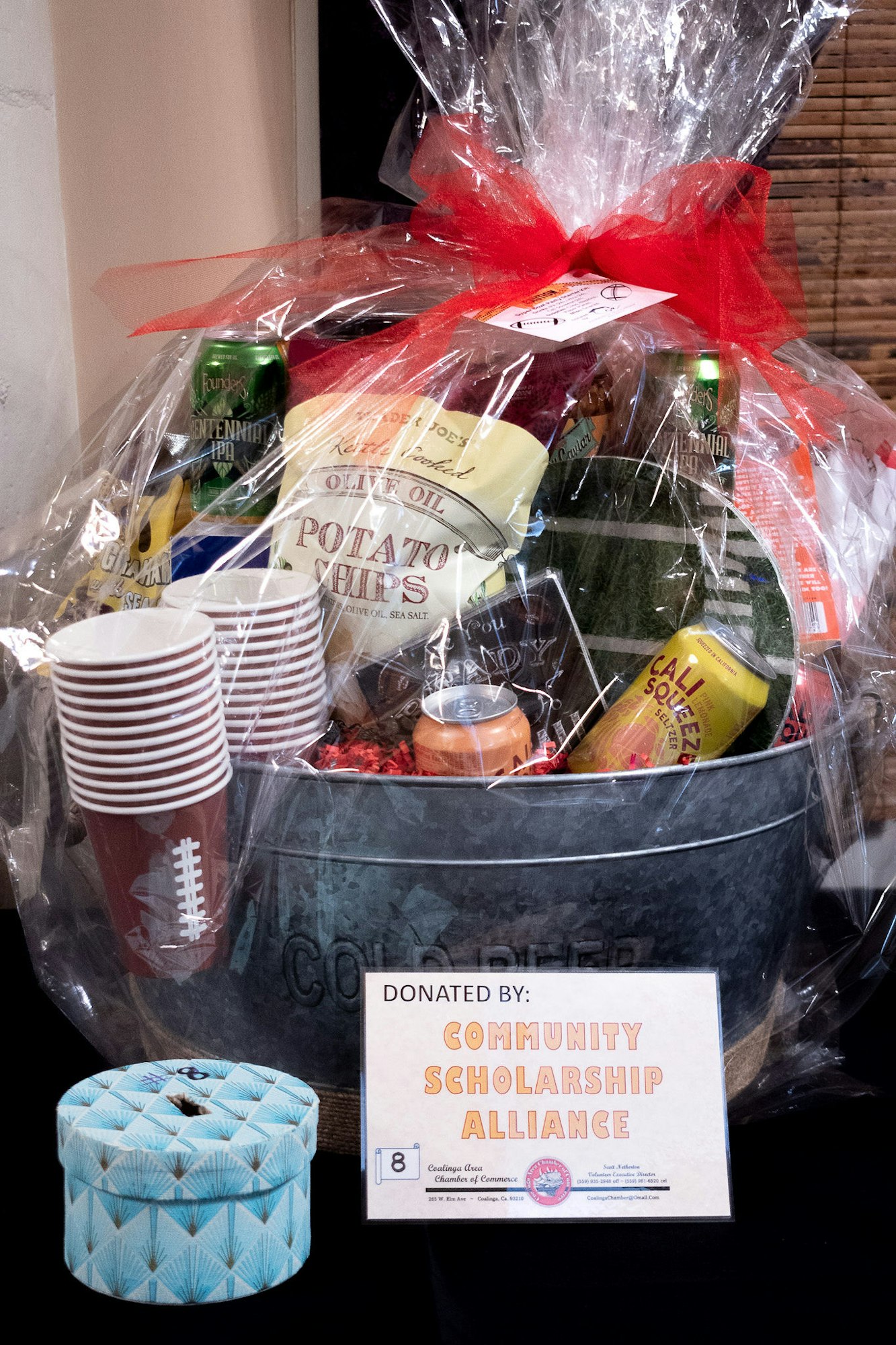 A gift basket with various items including beer, chips, and olive oil, wrapped in clear plastic with a red bow, donated by a community group.