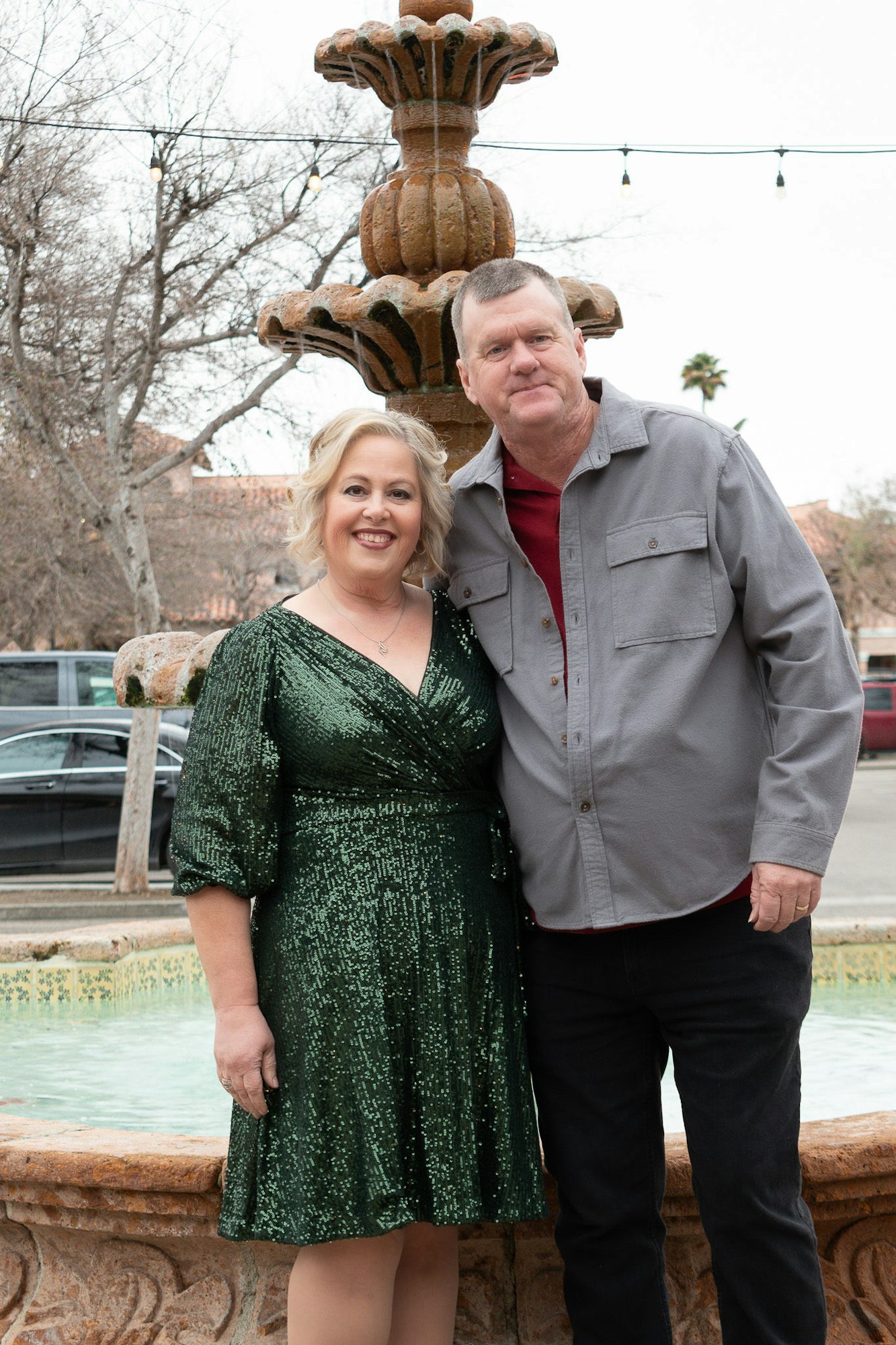 Two people posing in front of a fountain with string lights and trees in the background.