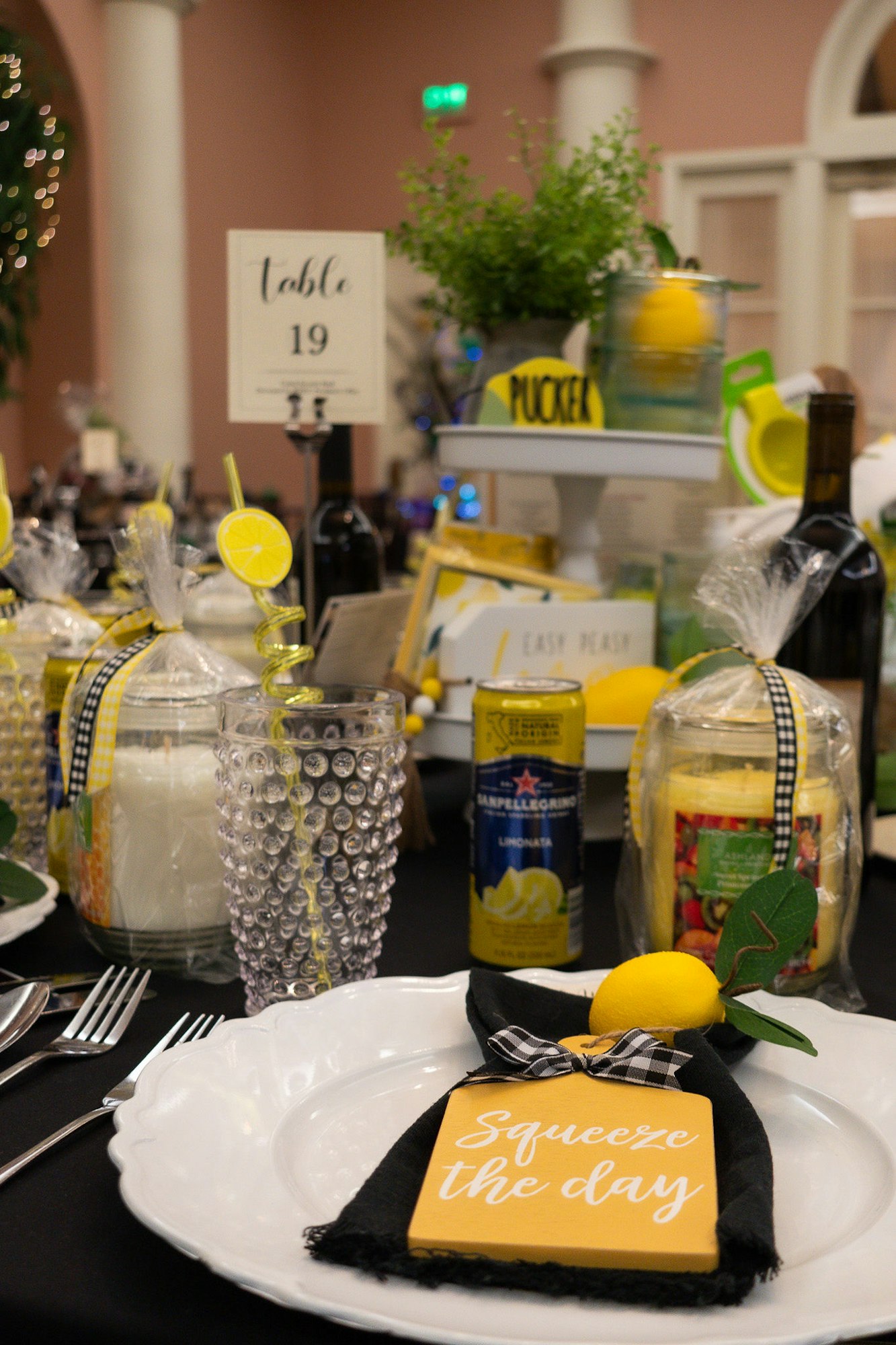 Lemon-themed table setting with a "Squeeze the day" card, beverages, potted plant, and a table number sign.