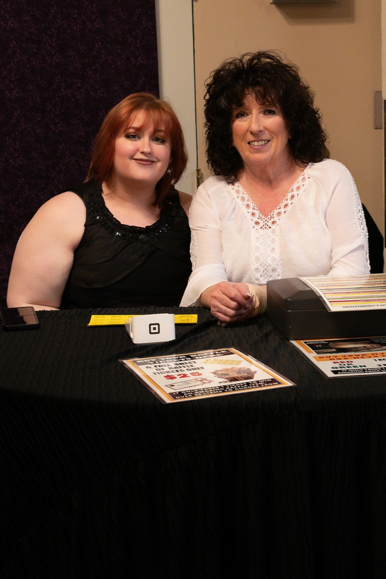 Two women smiling at a table with flyers, a card reader, and a phone.
