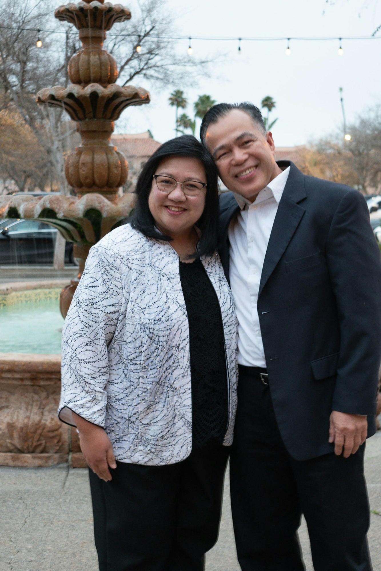 Two smiling people posing in front of a fountain, with festive lights in the background.