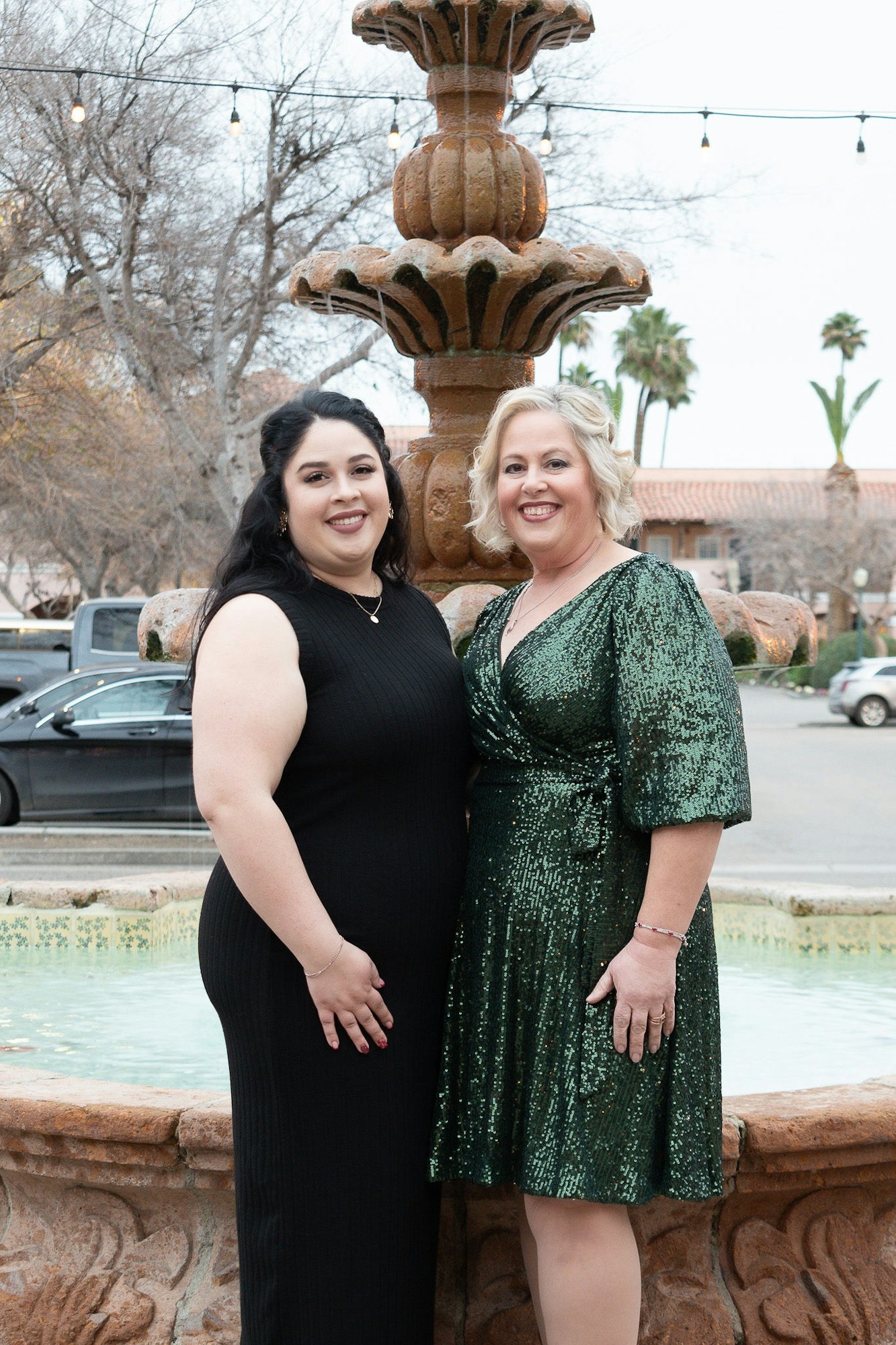 Two women in dresses posing by a fountain, with string lights and palm trees in the background.