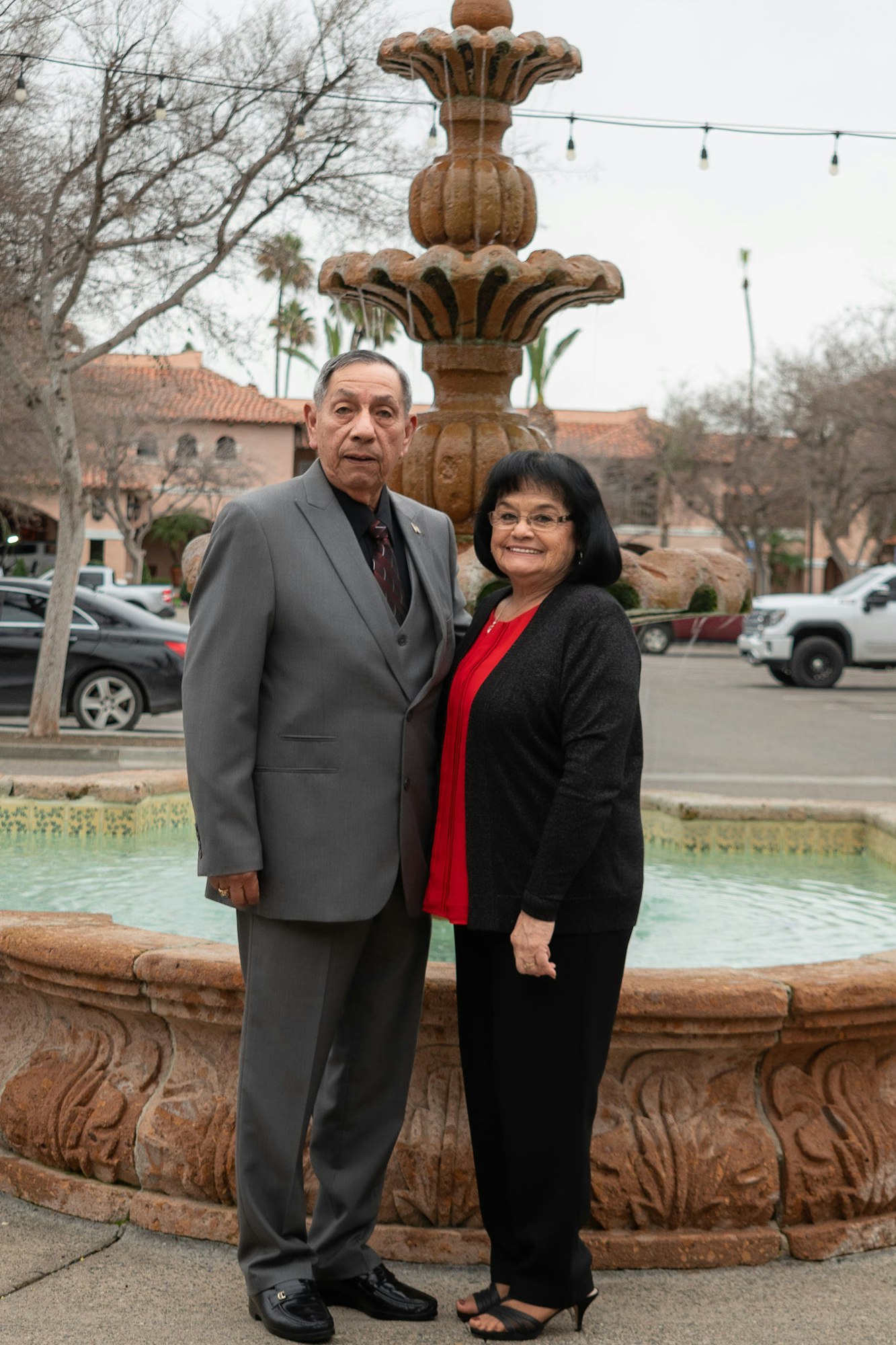 A couple standing in front of a fountain, smiling, dressed in smart attire.