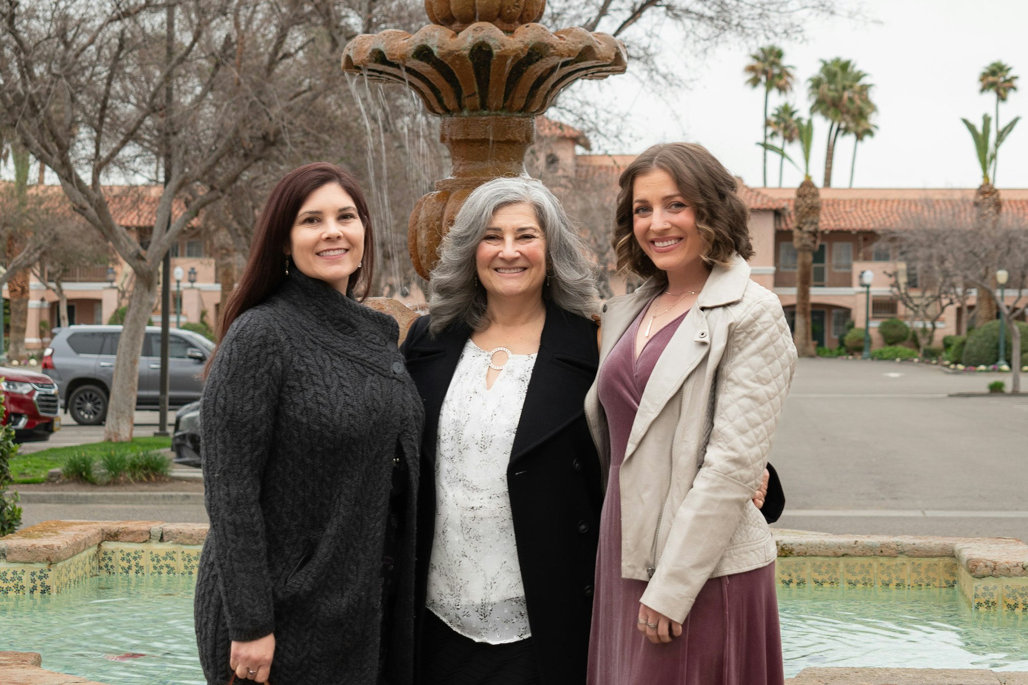 Three women smiling in front of a fountain with palm trees and a building in the background.