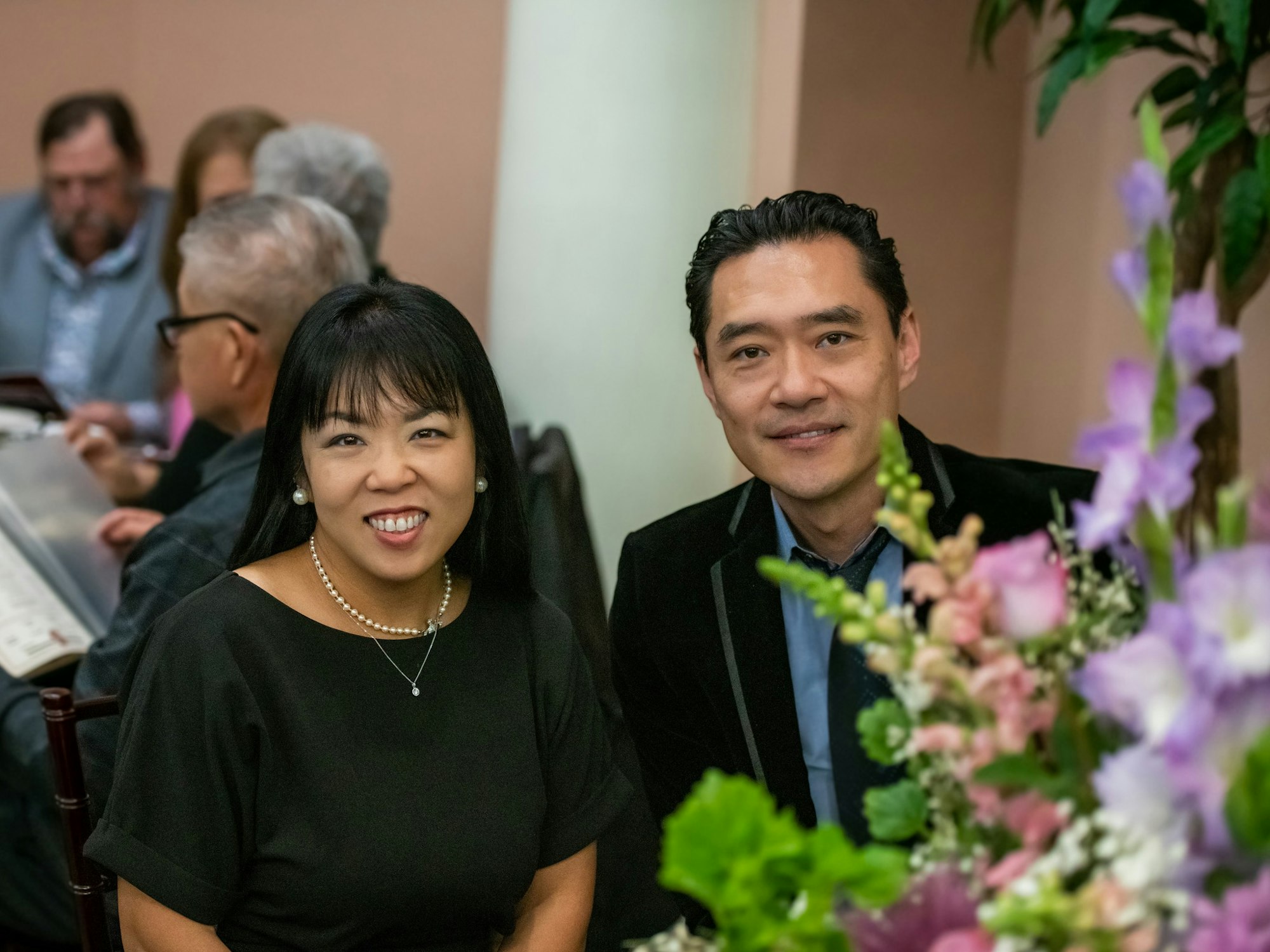 May contain: flower, flower arrangement, plant, flower bouquet, people, person, clothing, formal wear, suit, accessories, jewelry, necklace, face, head, photography, portrait, blazer, coat, jacket, adult, male, man, female, woman, purple, glasses, tuxedo, potted plant, and dress