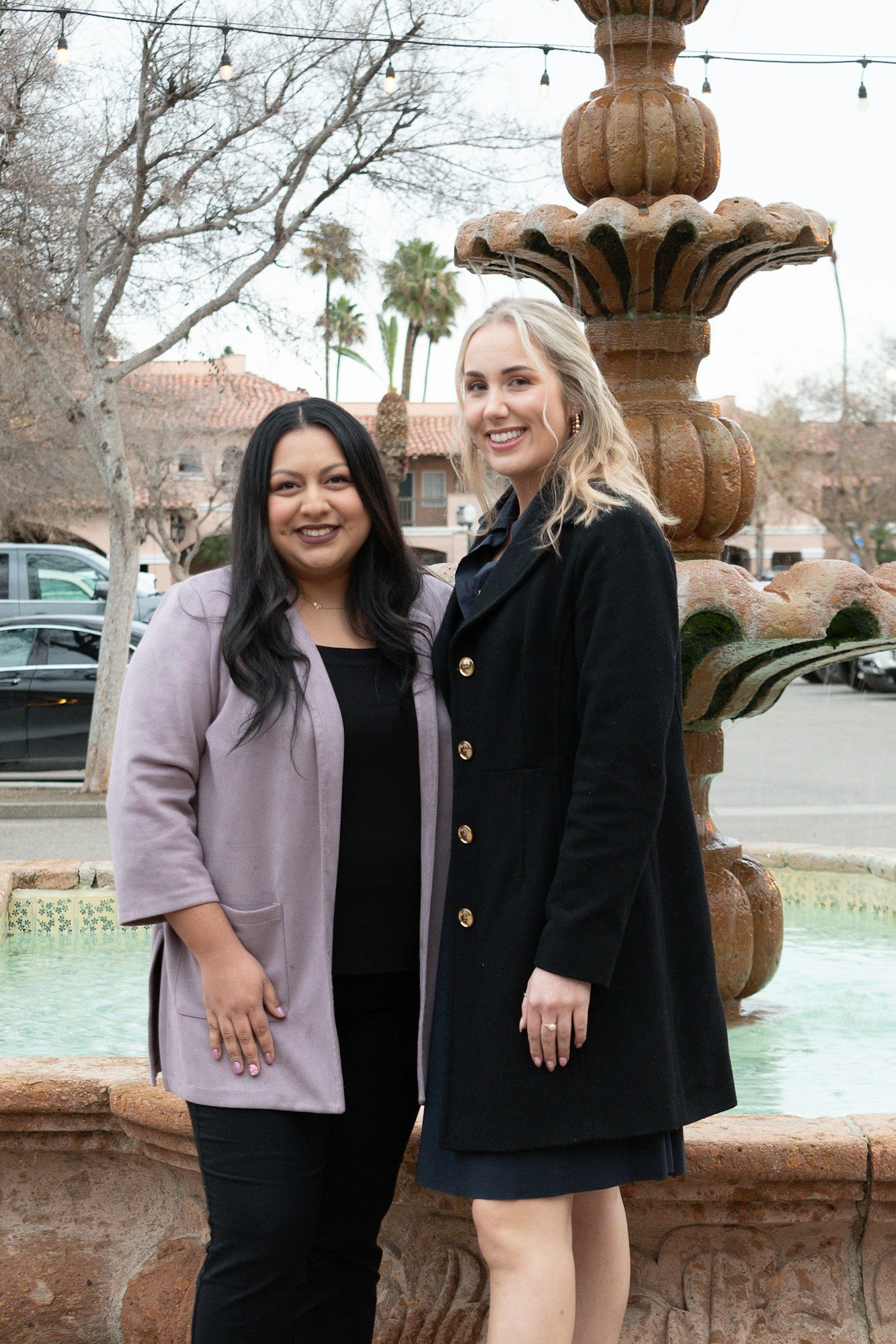 Two women smiling by a fountain, with trees and string lights in the background.