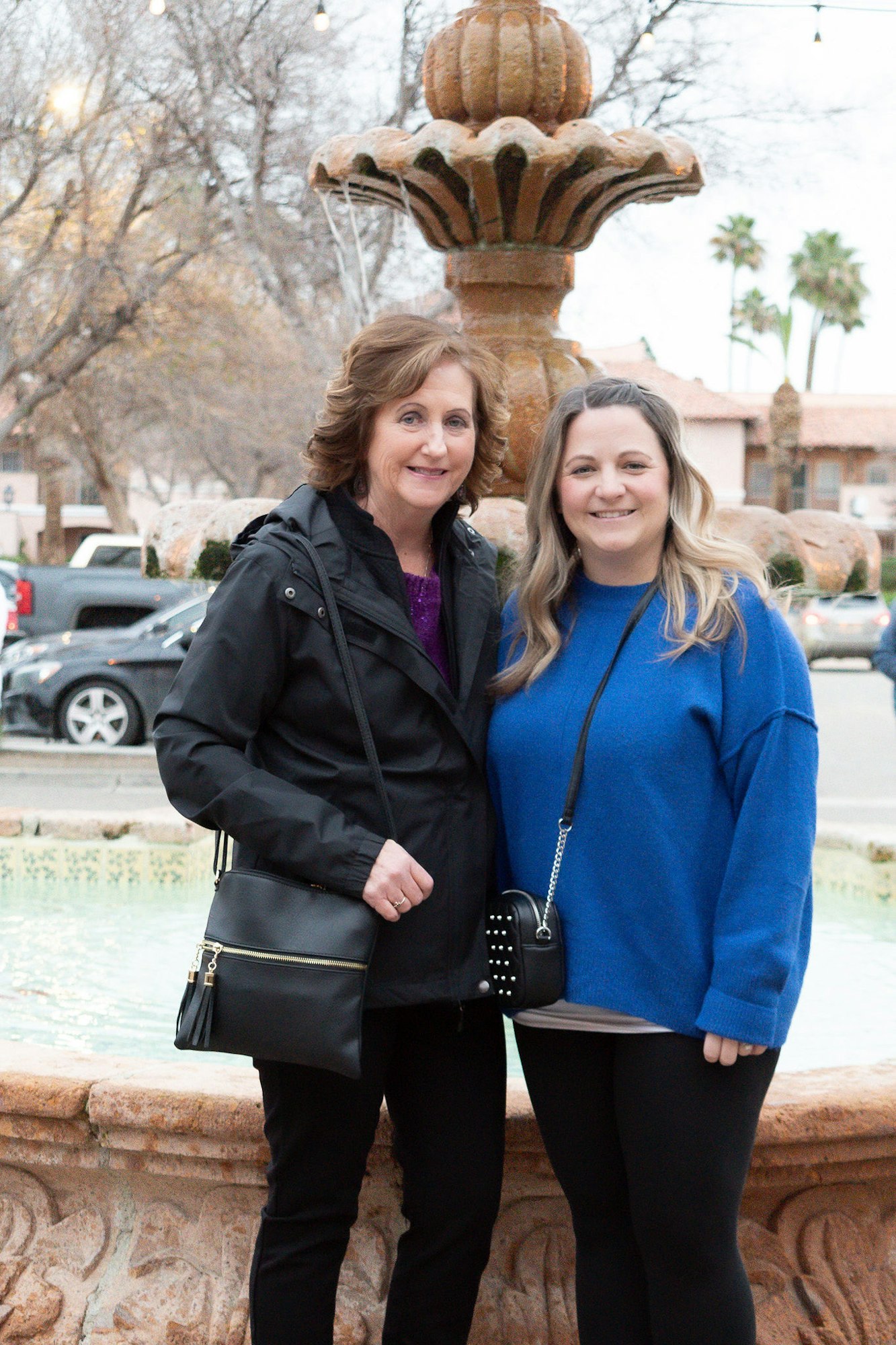 Two women smiling in front of a fountain, with trees and parked cars in the background.