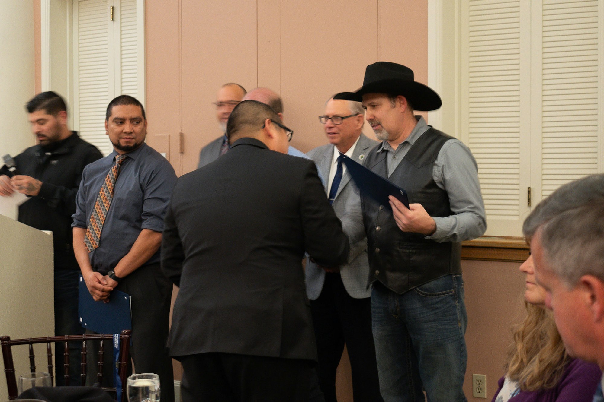 Men in a meeting room, one wearing a cowboy hat, appear to be in a discussion or presentation.