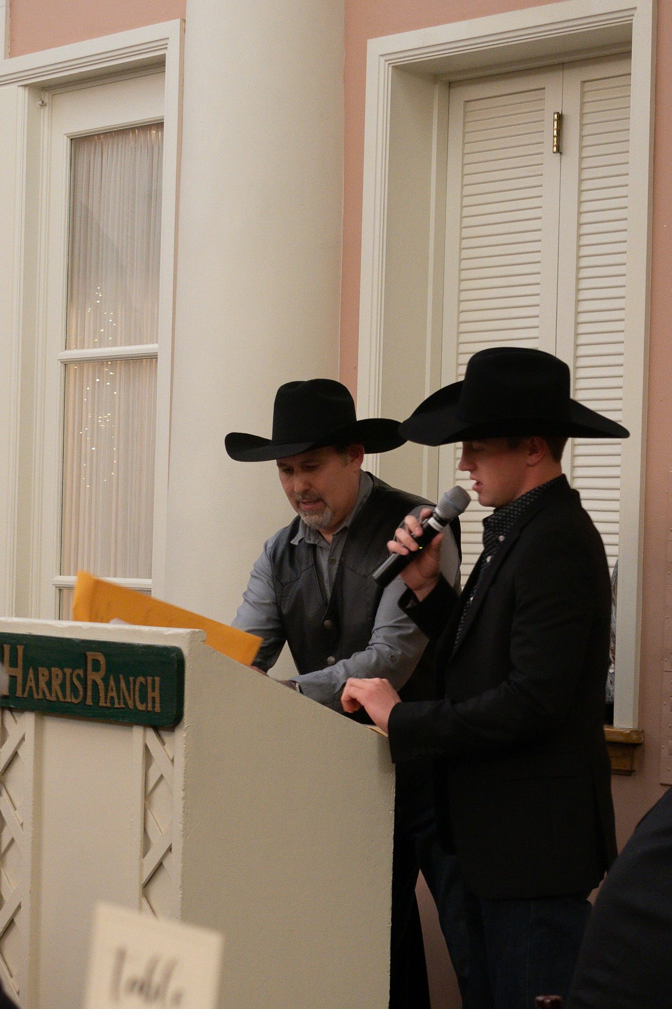 Two people in cowboy hats speaking at a podium with a sign "Harris Ranch."