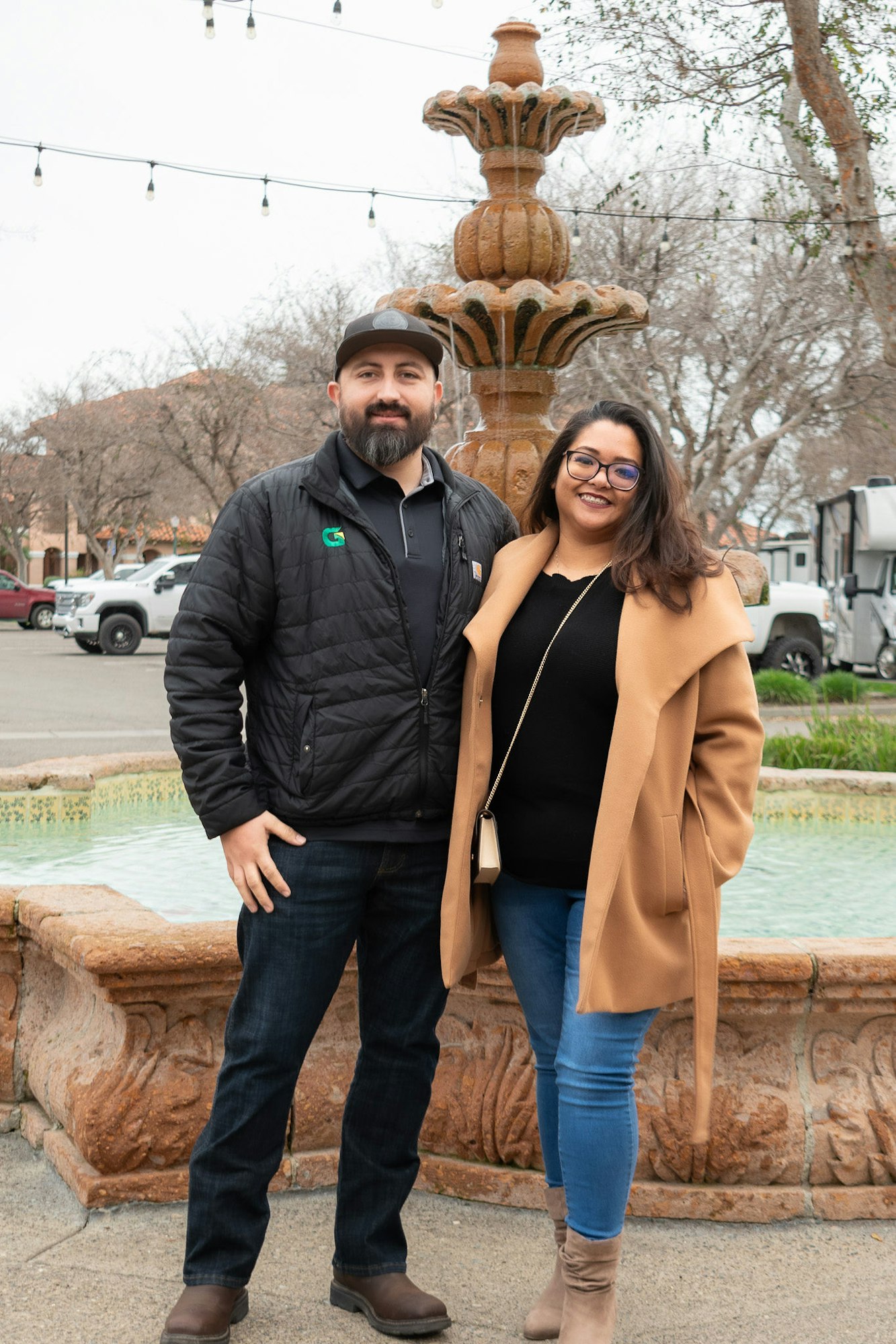 A man and a woman smiling in front of a water fountain, with string lights and trees in the background.