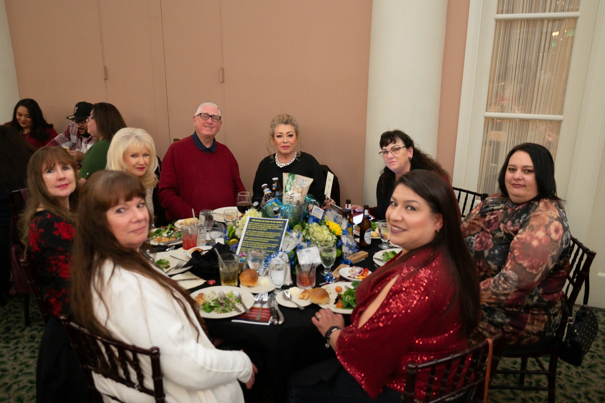 A group of people dining at a table with food and drinks, smiling at the camera.