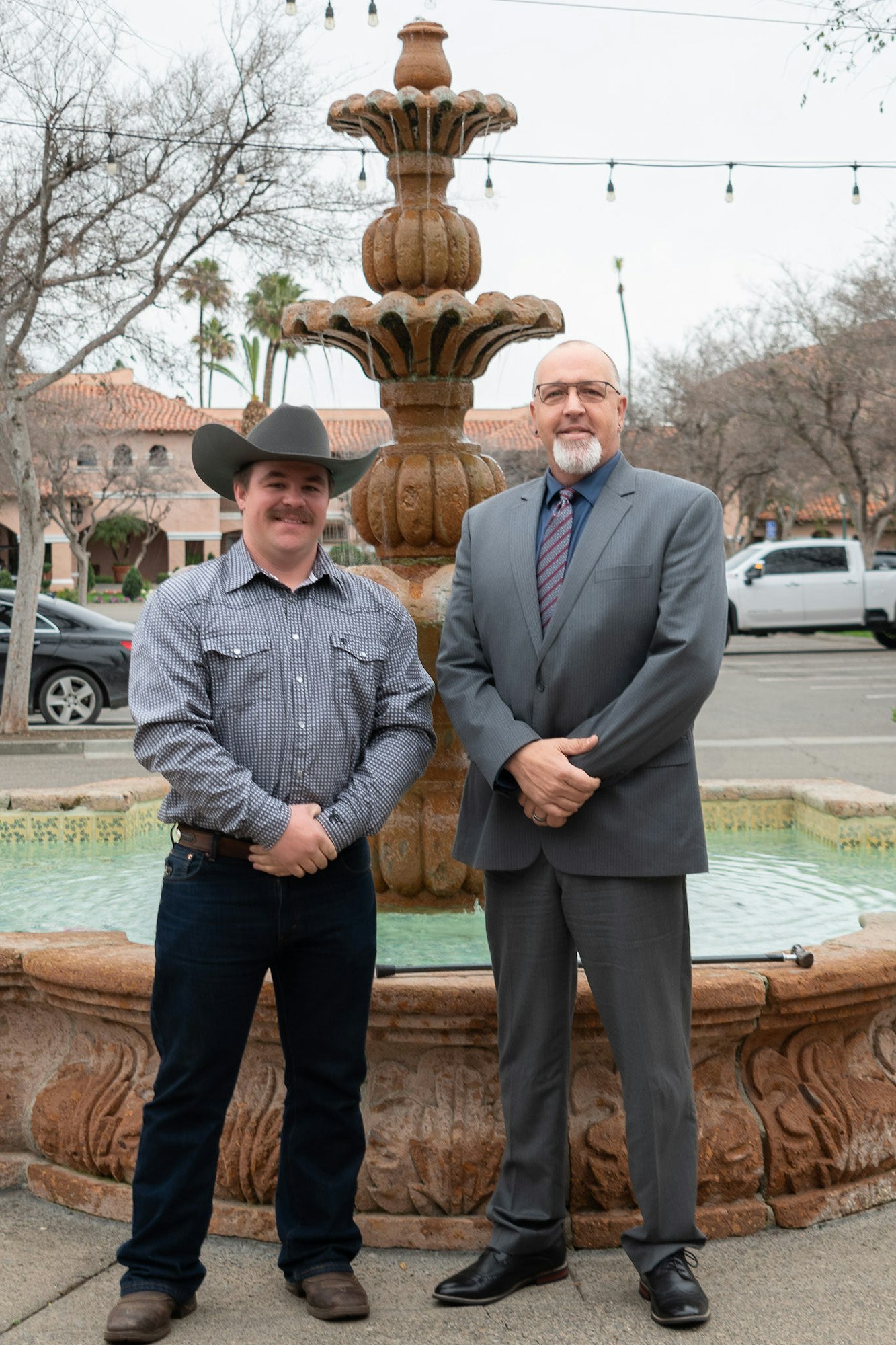 Two men in professional attire standing in front of a fountain with trees and string lights in the background.