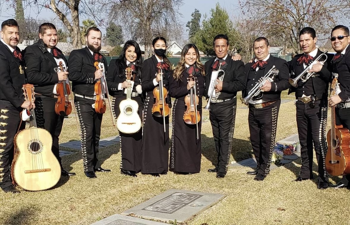 A mariachi group poses in traditional attire at a cemetery, holding instruments and celebrating together.