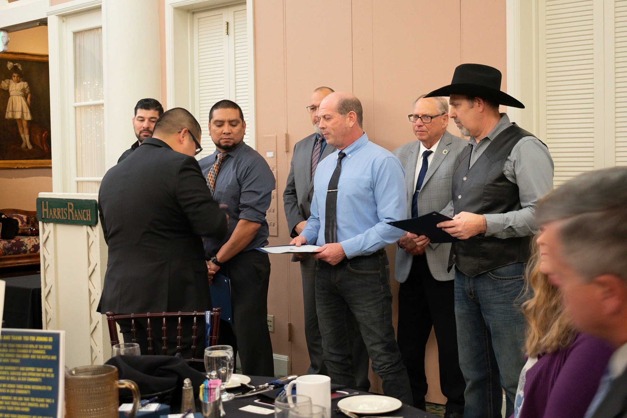 Men in business attire in a room, one receiving a document, others observing, a table set for a meal in the foreground.