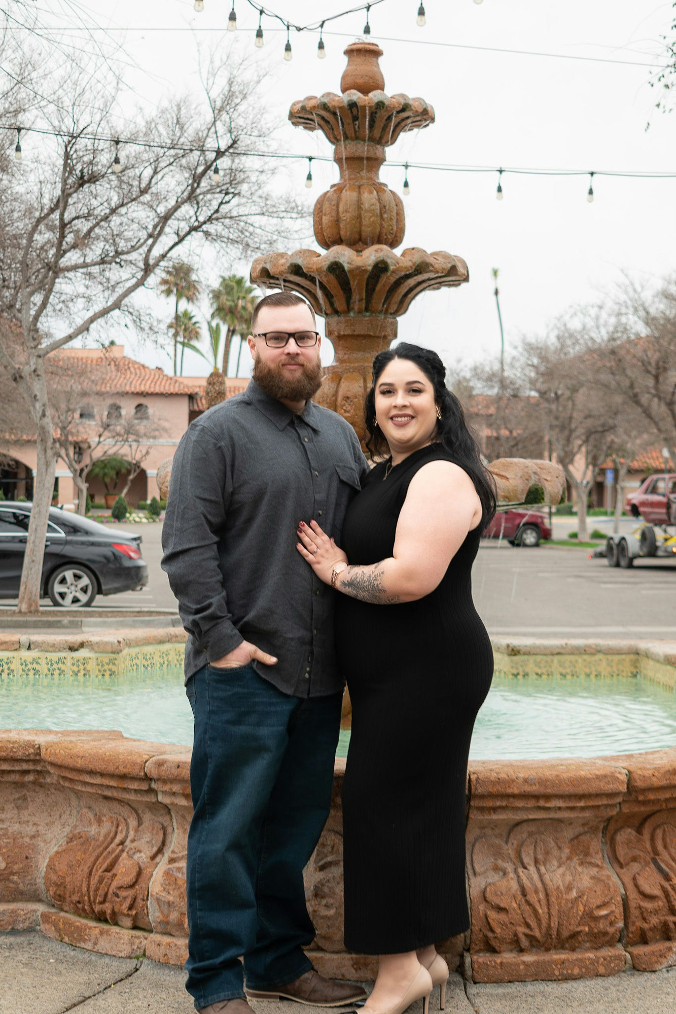 A couple posing in front of a fountain with string lights overhead.