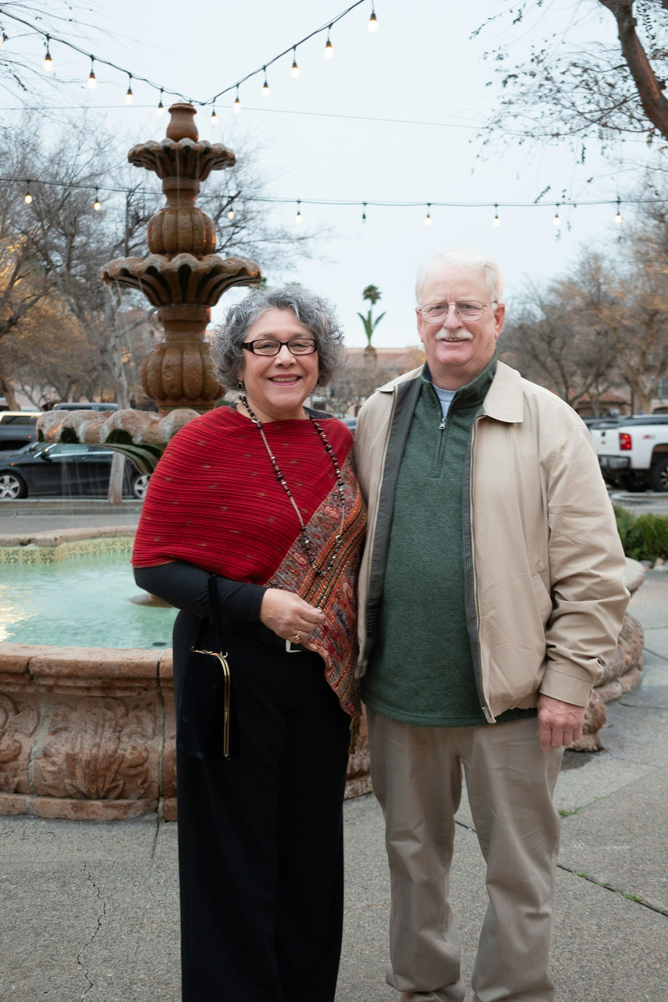 A smiling couple posing in front of a fountain with string lights.