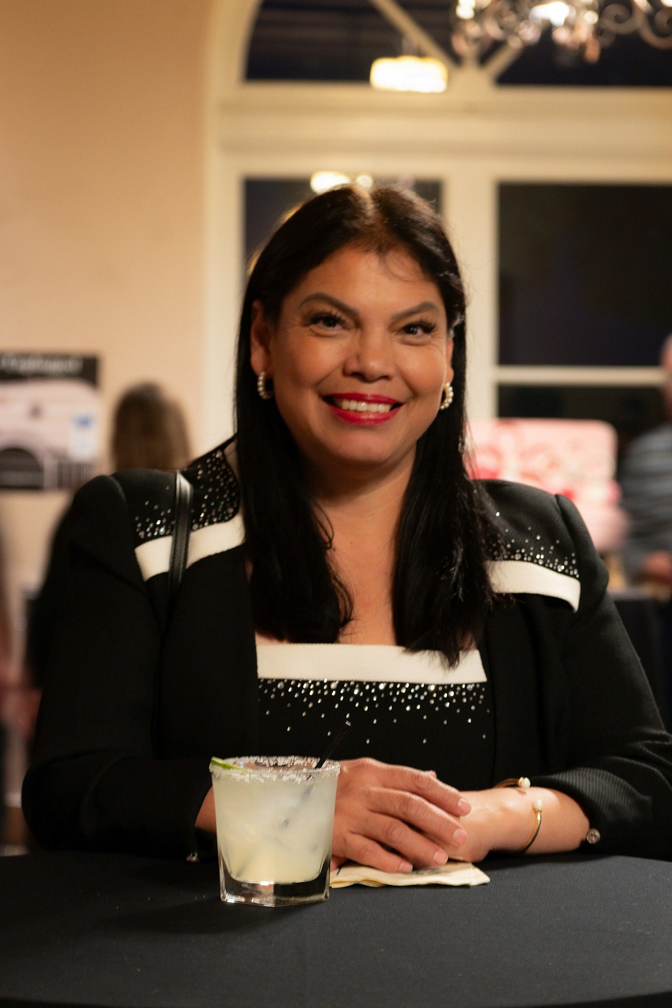 A smiling woman sitting with a cocktail in a social setting.