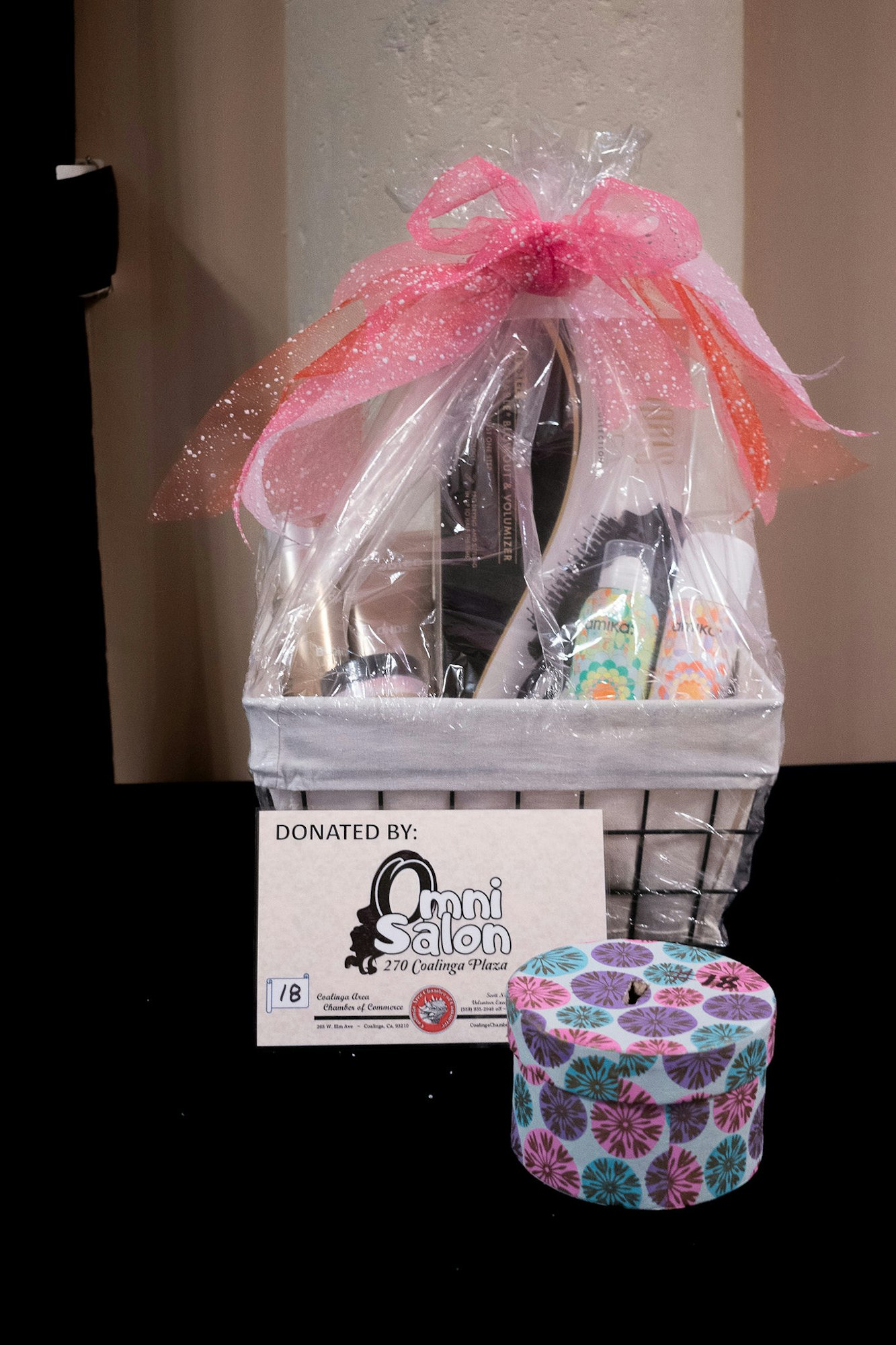 Gift basket with clear wrapping and pink bow, sign indicating donation by a salon, and a patterned box beside it.