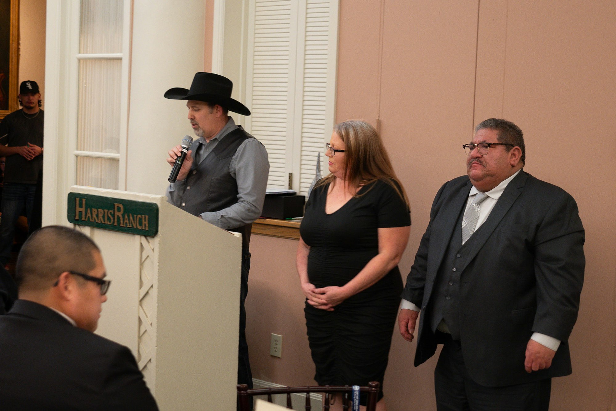 Man in a cowboy hat speaking at a podium, two people standing beside him, indoor setting.