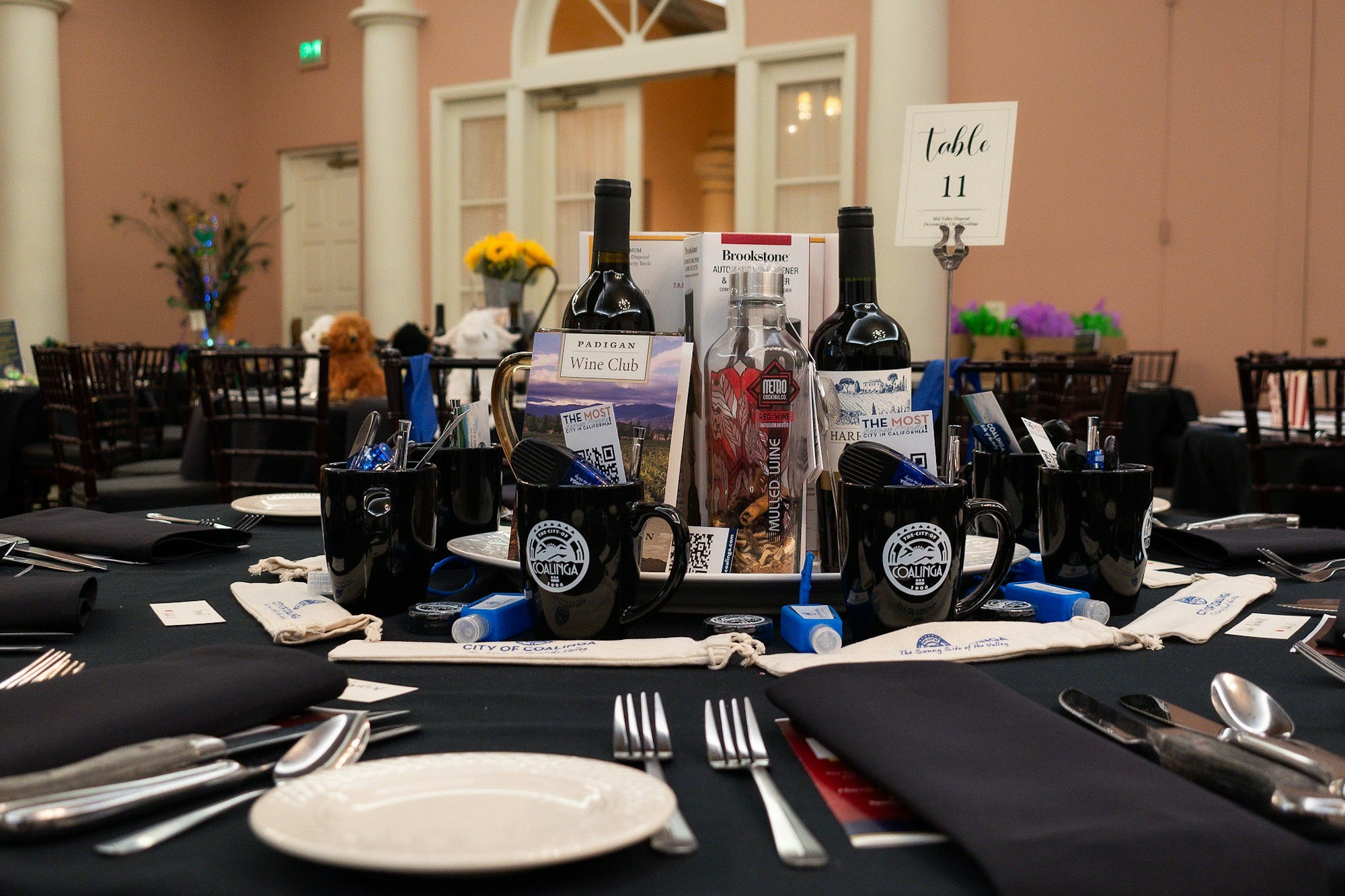 A decorated banquet table with wine bottles, mugs, a brochure, and a "Table 11" sign; event setting in the background.