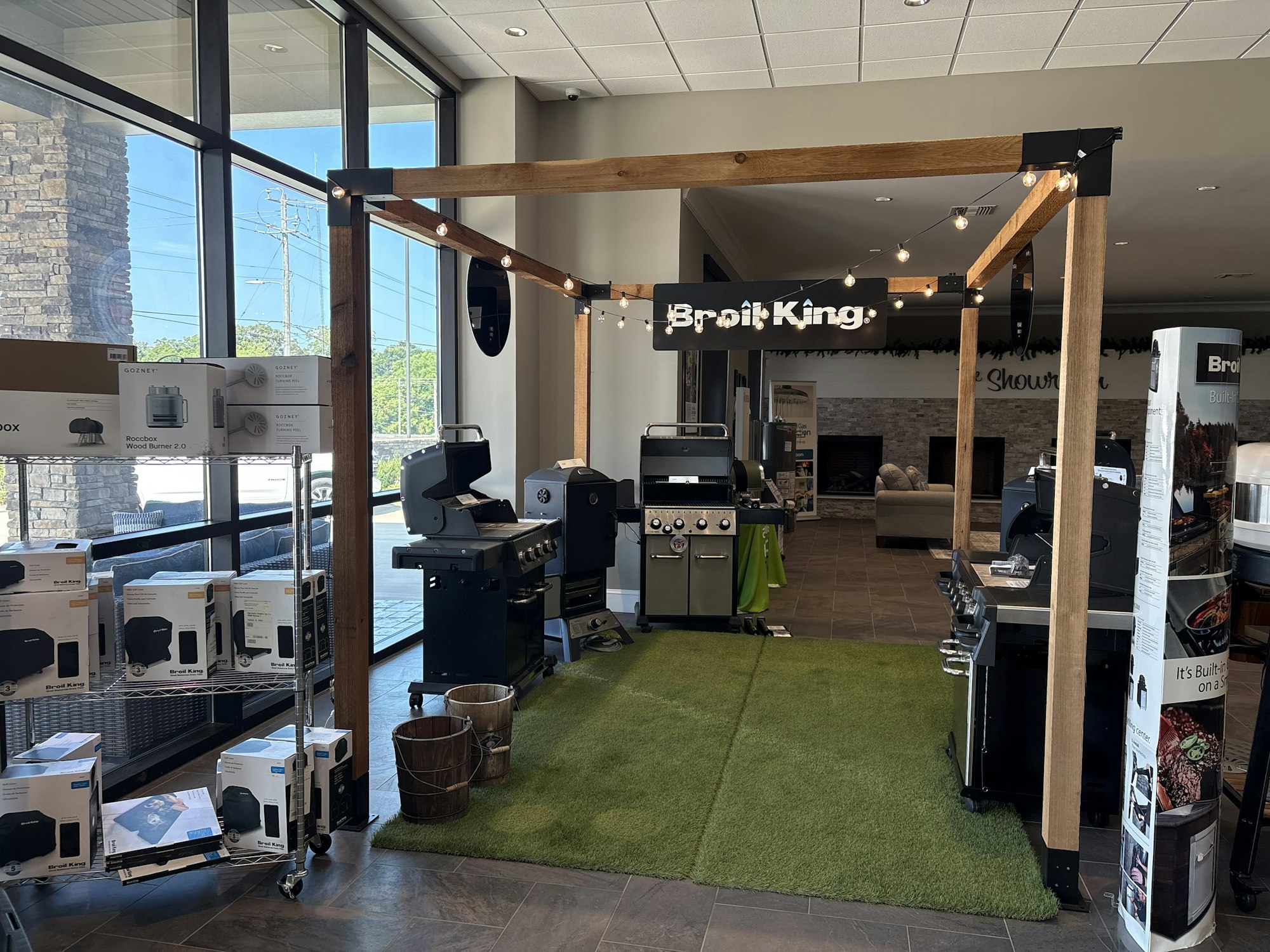 The image shows a display area for Broil King grills, featuring multiple grill units and related products in a store setting.