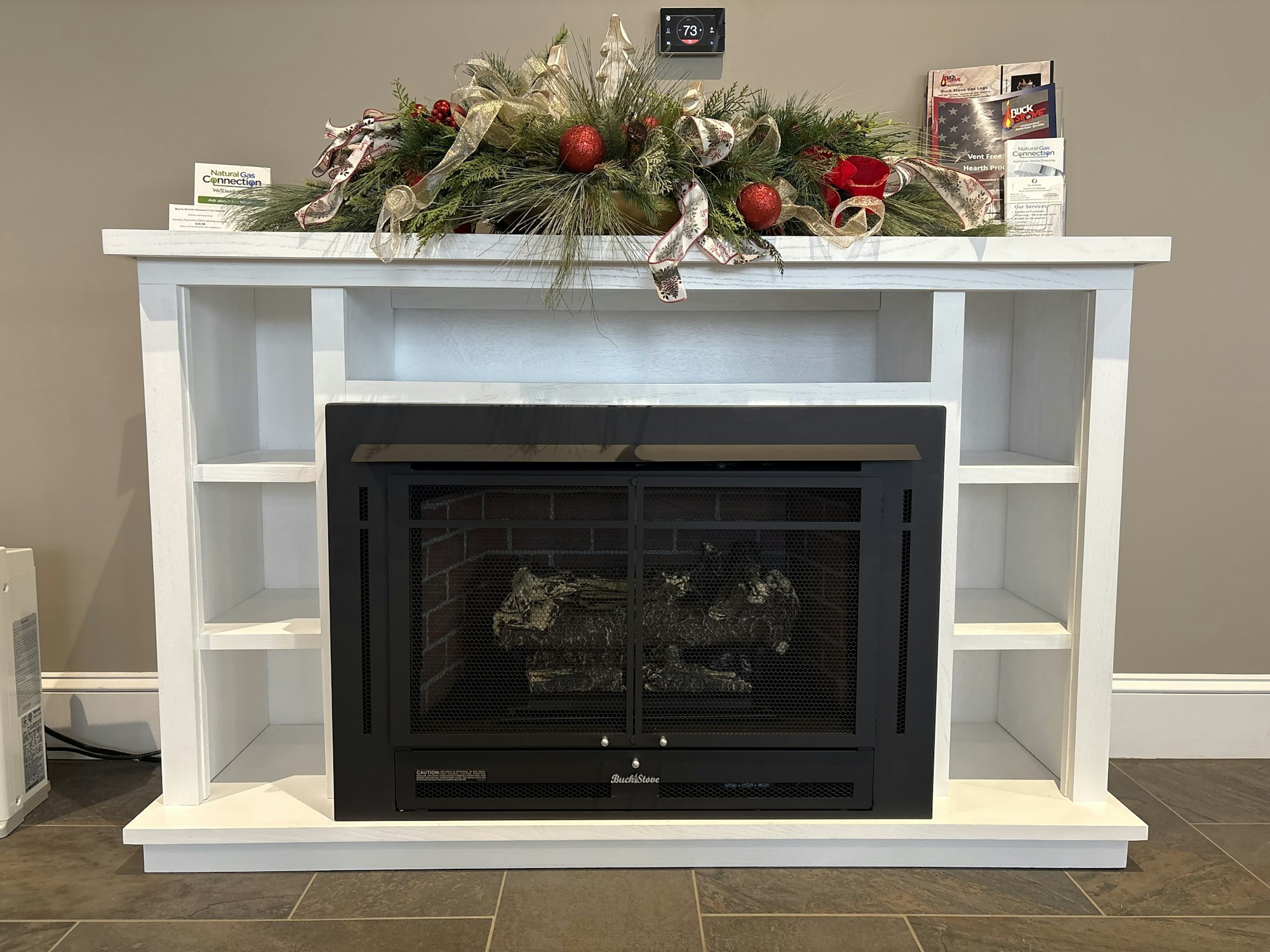 A decorated white fireplace with shelves and a festive arrangement of greenery and ornaments on top.