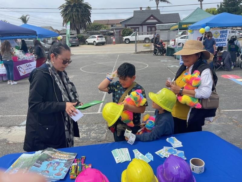 A woman talks to two kids holding colorful plush toys and yellow helmets at an outdoor event with booths and tents.