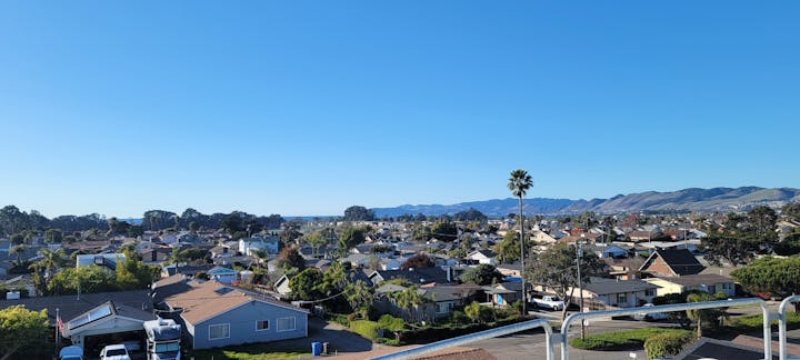 Suburban neighborhood with houses, greenery, and a clear blue sky; hills in the distance.