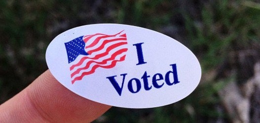 An "I Voted" sticker featuring a U.S. flag, held between fingers.