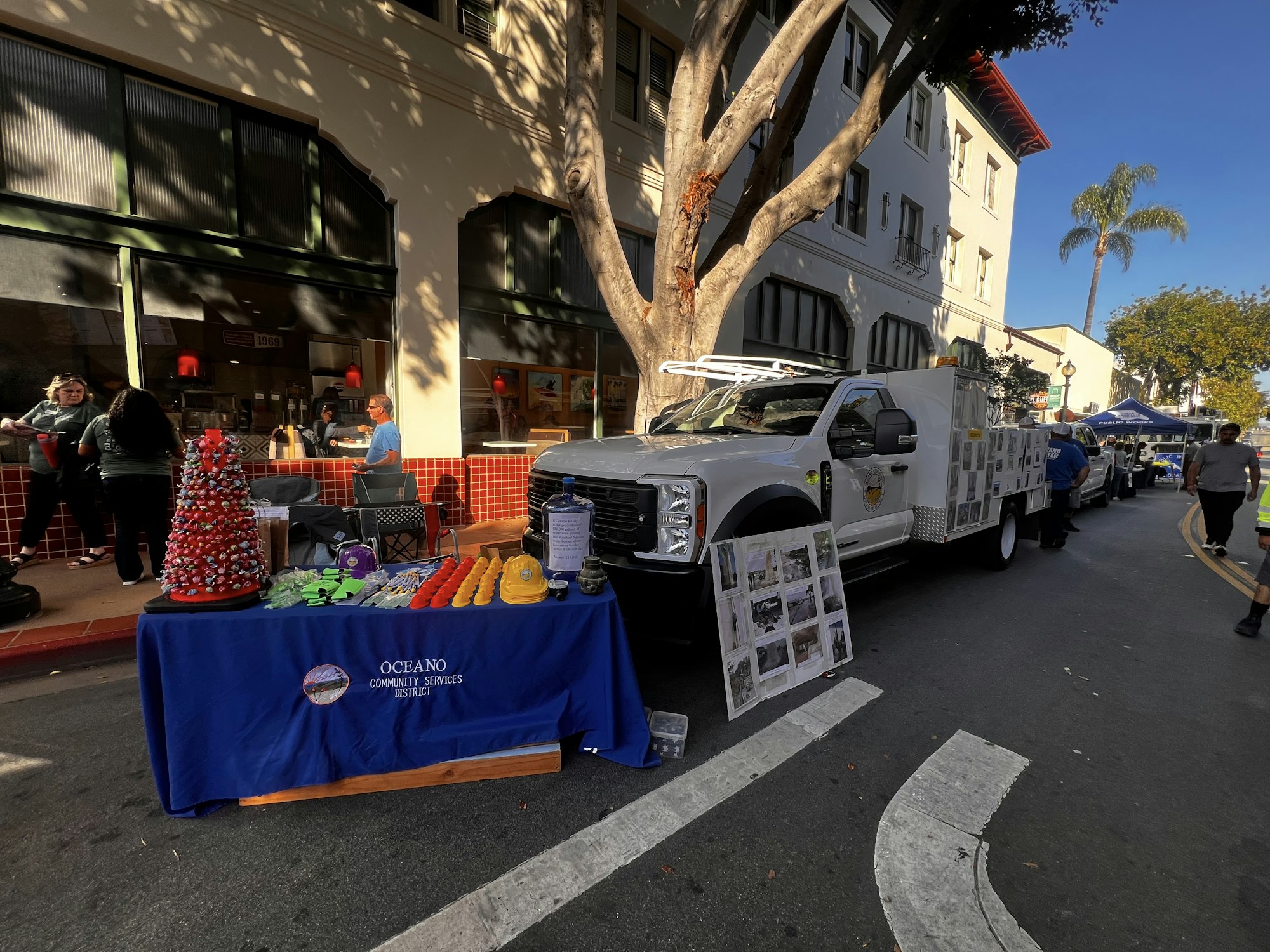 A street scene featuring a vendor table with promotional items, a white truck, and people interacting outside a building.